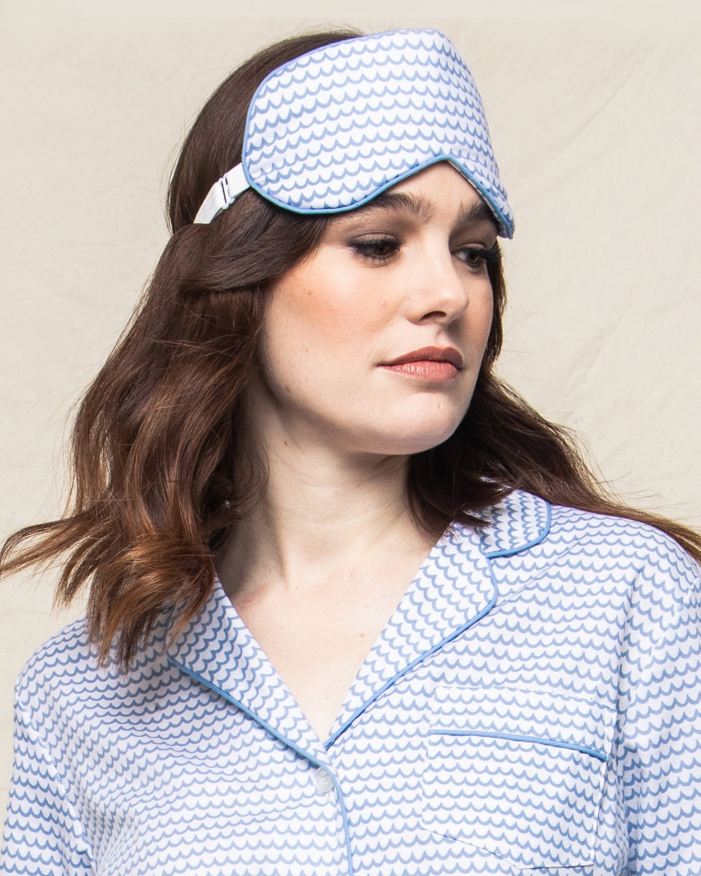 A brown-haired woman models luxury Petite Plume sleepwear, featuring the Adults Sleep Mask in La Mer and coordinating pajamas, against a neutral backdrop.