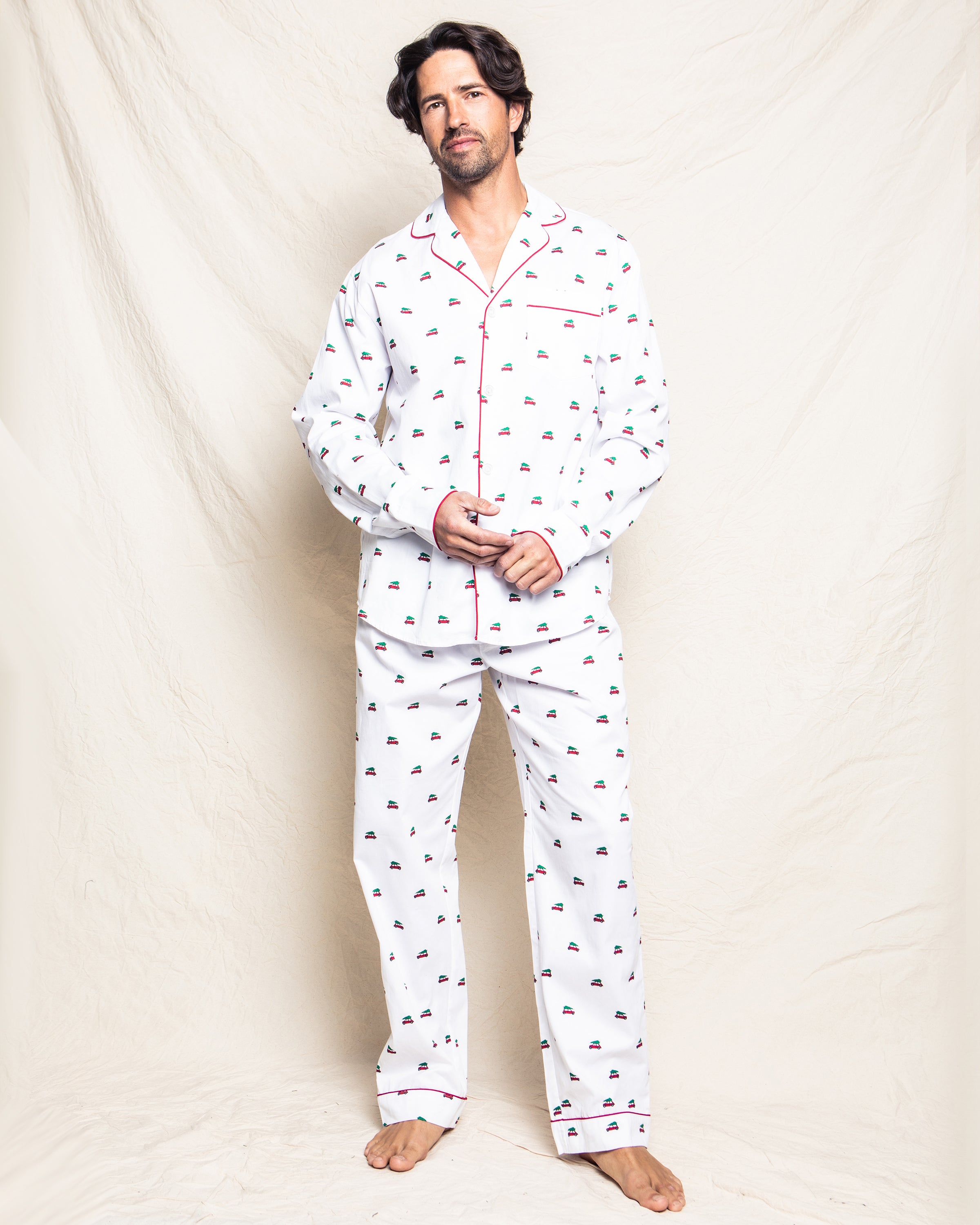 A man with dark hair stands barefoot against a light fabric backdrop, wearing the Petite Plume Mens Twill Pajama Set in Holiday Journey, featuring soft cotton twill with boat prints and red piping for a relaxed look.