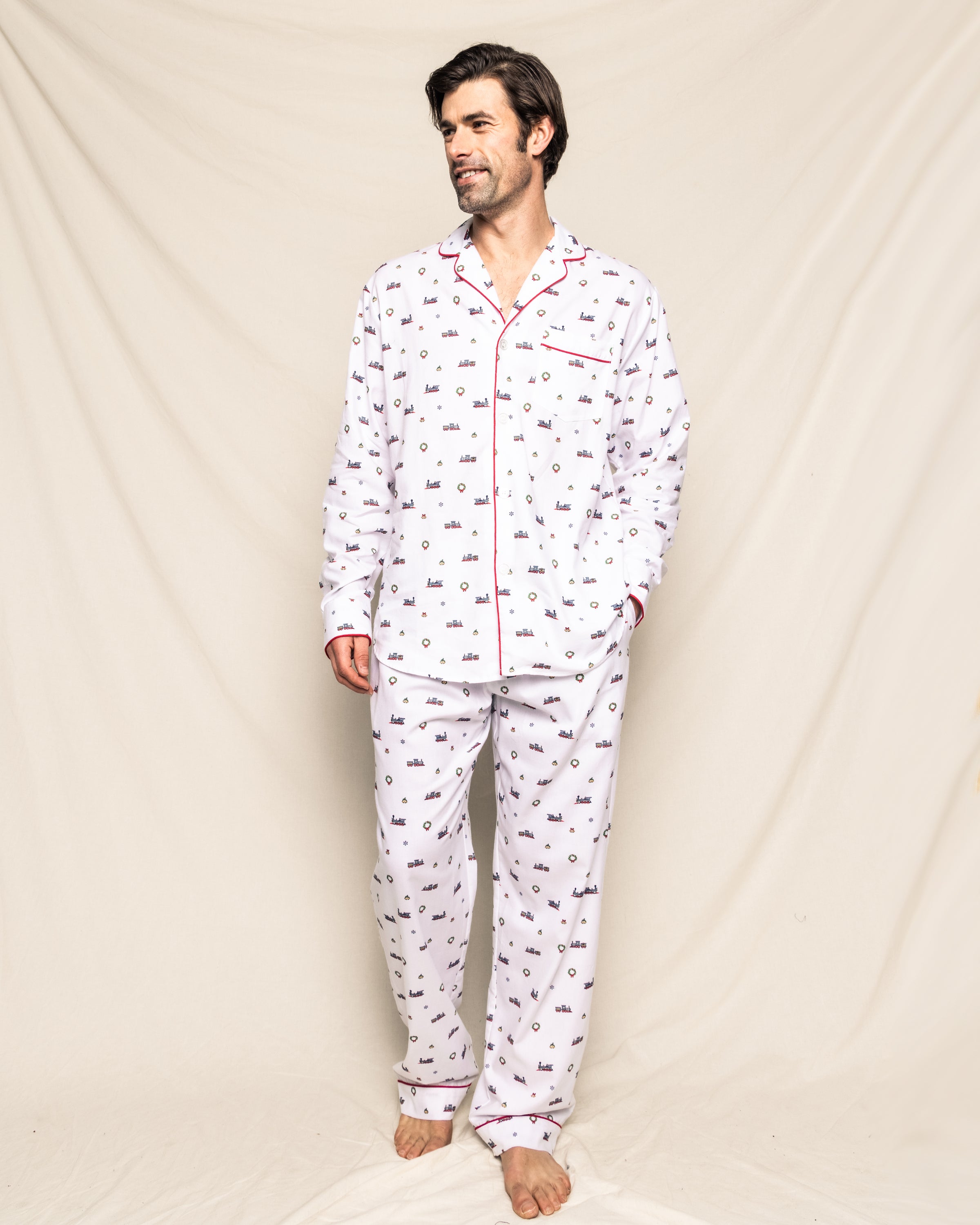A man stands barefoot against a cream background wearing Petite Plumes Mens Twill Pajama Set in Arctic Express, crafted from high-quality cotton with a small patterned design and red piping. His hands rest by his sides as he gazes slightly to the right.