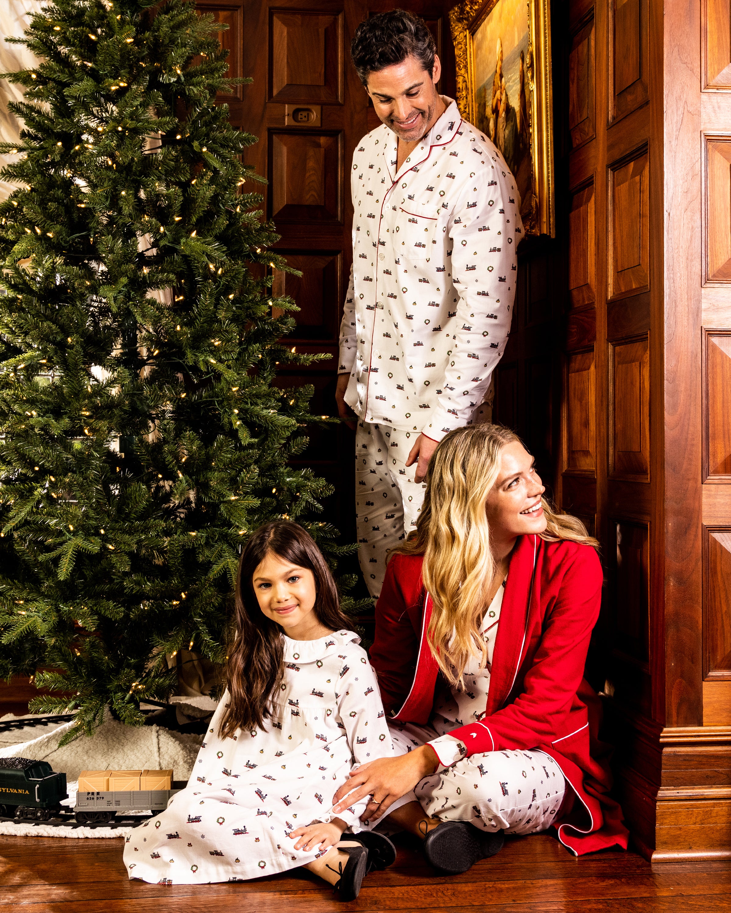 A man wearing a Petite Plume Mens Twill Pajama Set in Arctic Express stands by a Christmas tree with a woman in a red robe and a girl in cotton sleepwear, sitting joyfully among gift boxes beneath the warmly lit, wood-paneled room.