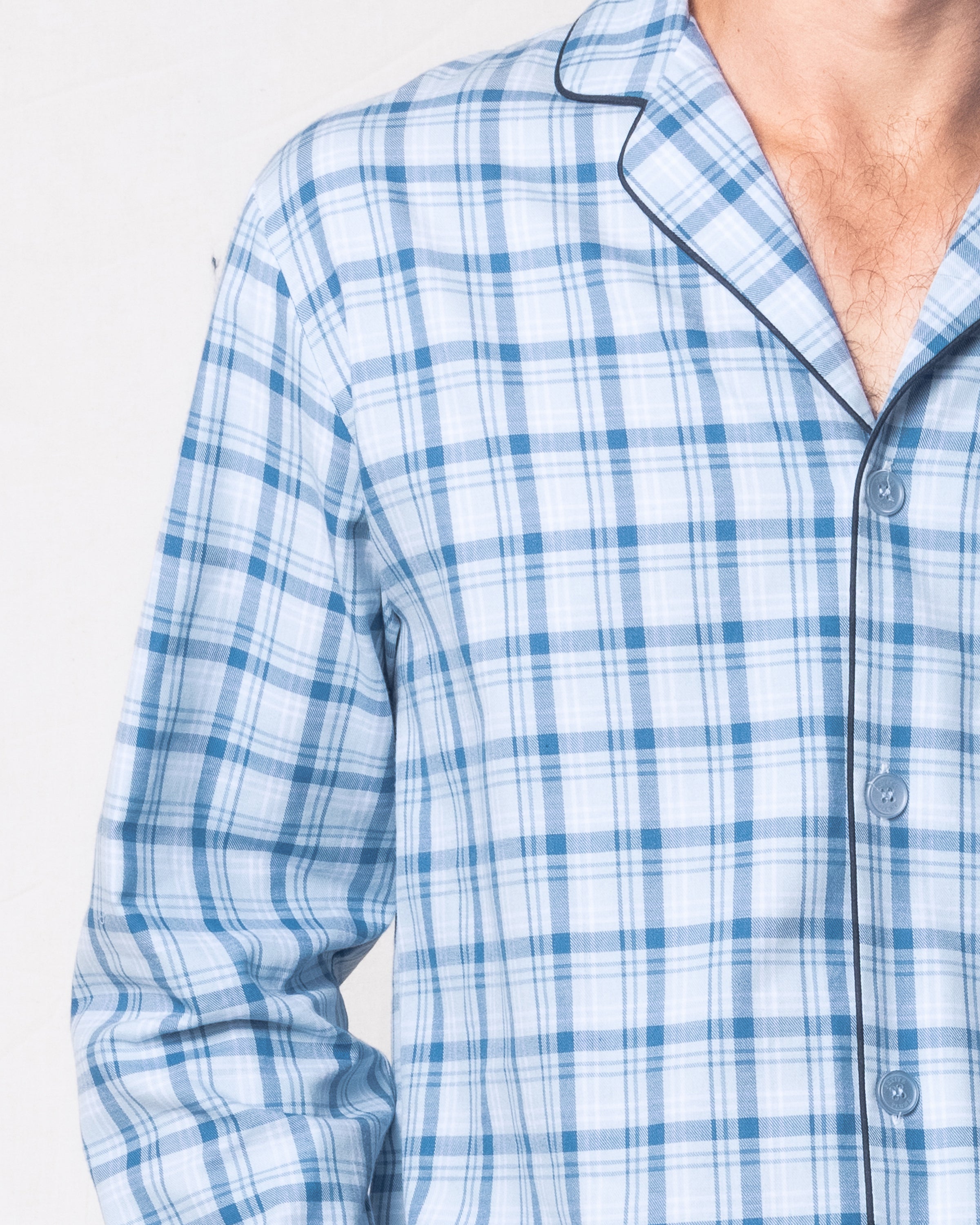 A person dons the Petite Plume Mens Twill Pajama Set in Seafarer Tartan, featuring blue and white plaid with black piping. The premium cotton shirt has buttons and a collar. The image focuses on the upper torso and partial arm against a plain backdrop.
