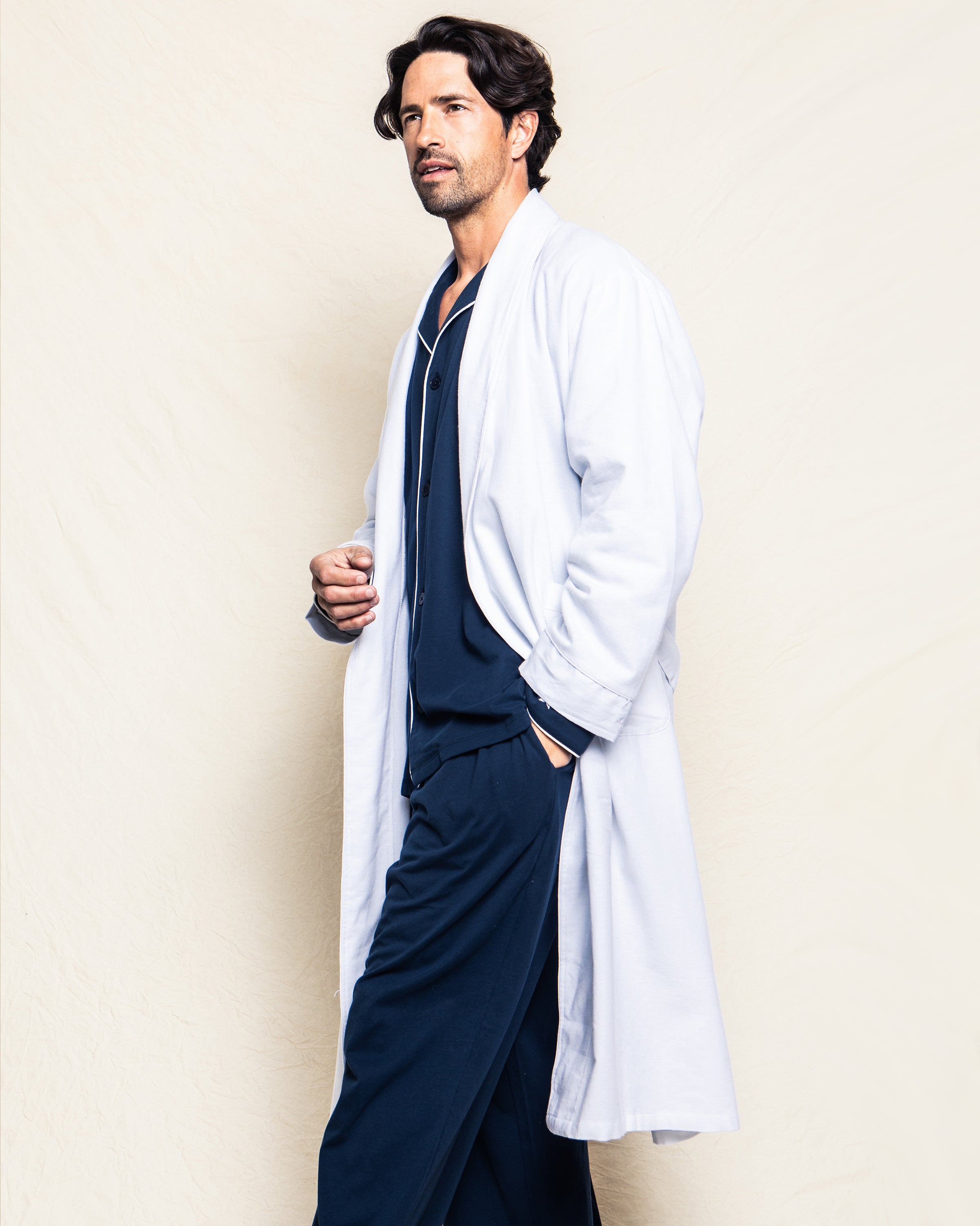 A man with wavy dark hair stands against a light backdrop wearing Petite Plumes luxury Mens Flannel Robe in white over navy blue 100% cotton pajamas, his left hand in his pocket as he gazes slightly to the side.