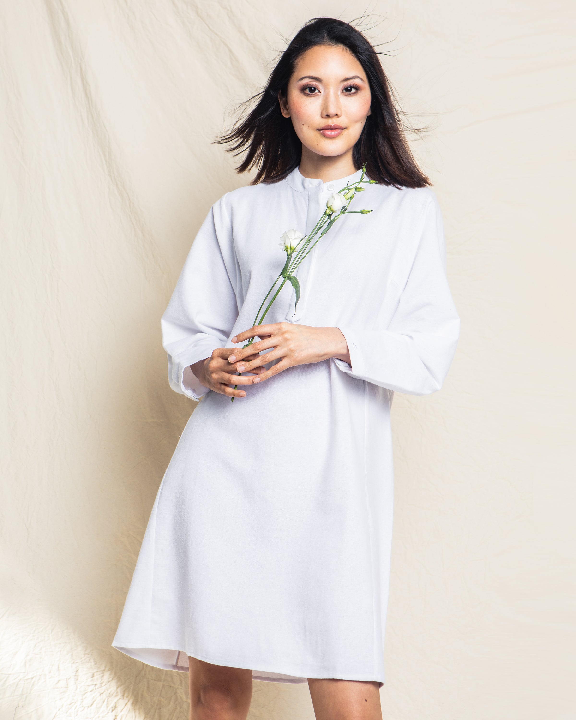 A person wears the Womens Flannel Grace Nightgown in White by Petite Plume, holding a white flower against a light beige backdrop. Their loose hair and confident gaze convey an elegant aura reminiscent of luxury sleepwear.