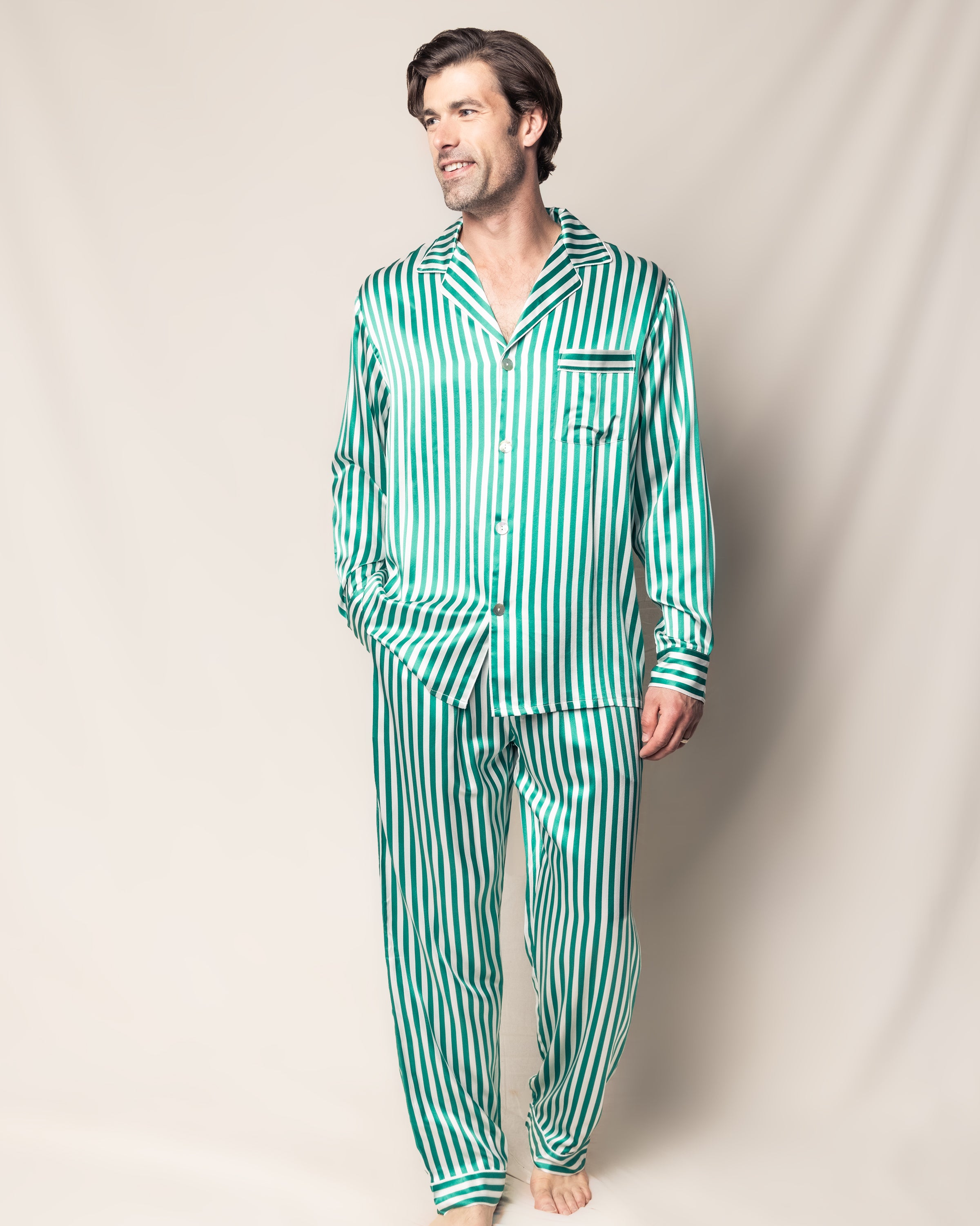 A man in a luxurious Petite Plume Mens Silk Pajama Set in Green Stripe stands barefoot against a neutral background, exuding elegance with a relaxed sideward glance.