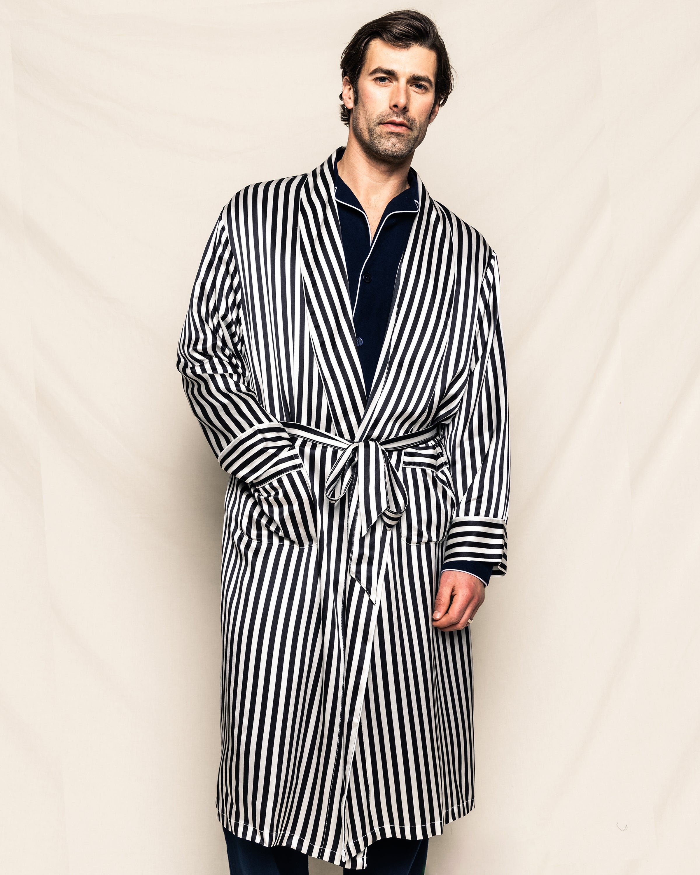 A man in a Petite Plume Mens Silk Long Robe in Bengal Stripe over a dark outfit stands against a plain background. He has short hair, a slight beard, hands partly in pockets, and wears a relaxed expression.