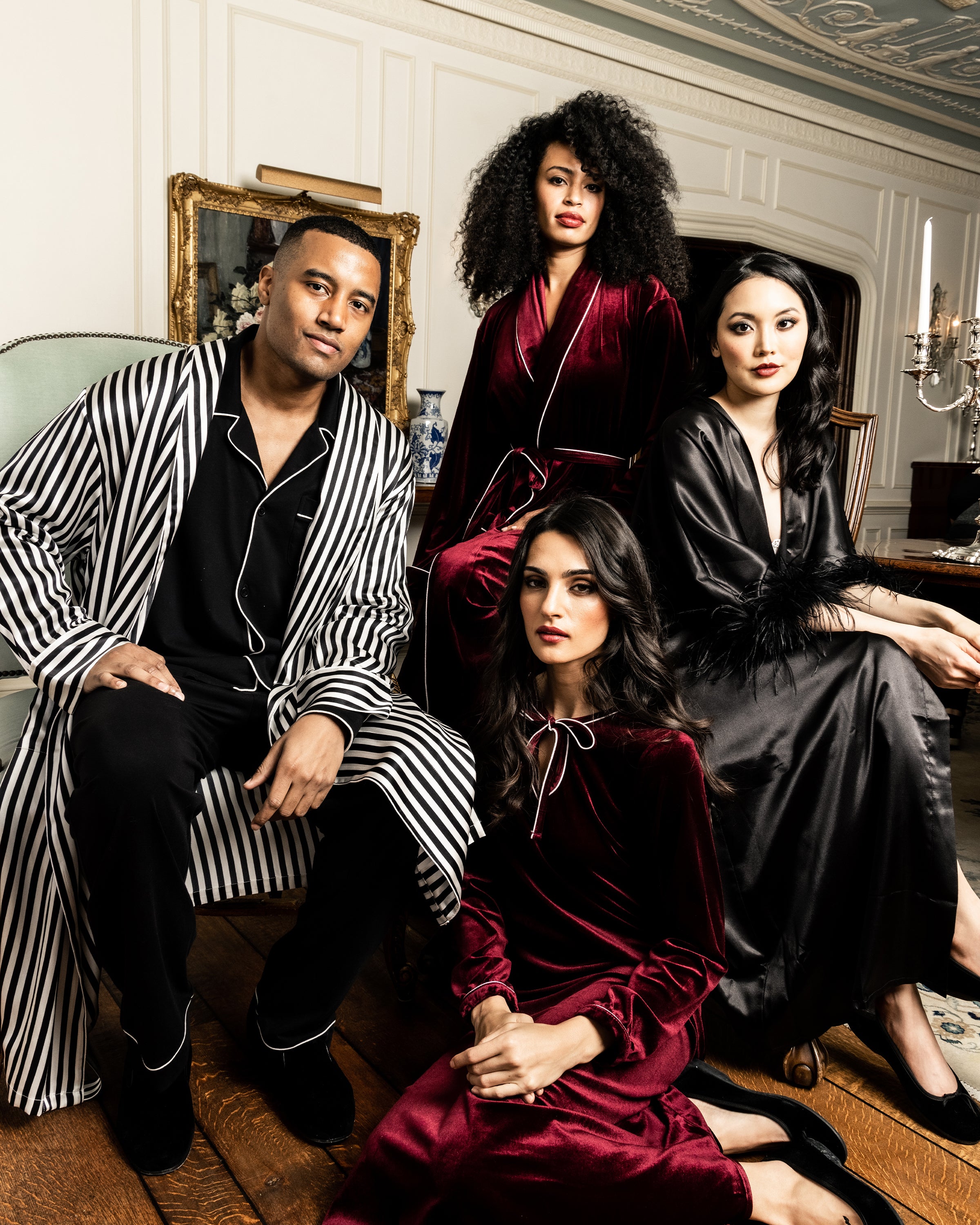 Four individuals in elegant Bengal Stripe silk robes by Petite Plume pose in a luxurious room with ornate decor. They sit and kneel on a wooden floor amidst a vintage chair, classic paintings, and a candle holder, exuding a sophisticated Hollywood style ambiance.