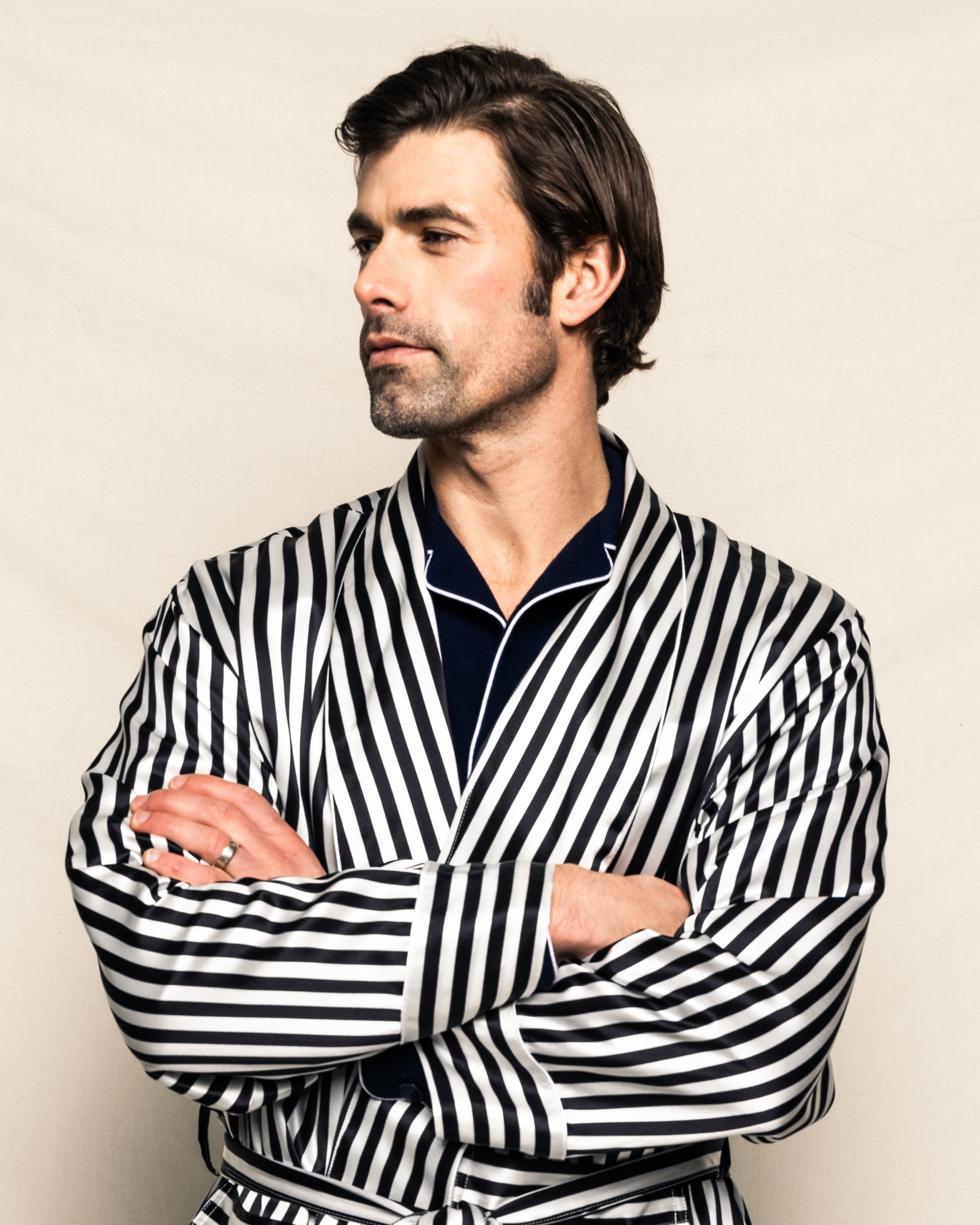 A man wearing Petite Plumes Mens Silk Long Robe in Bengal Stripe stands with arms crossed, looking to the side against a plain, light-colored fabric backdrop.