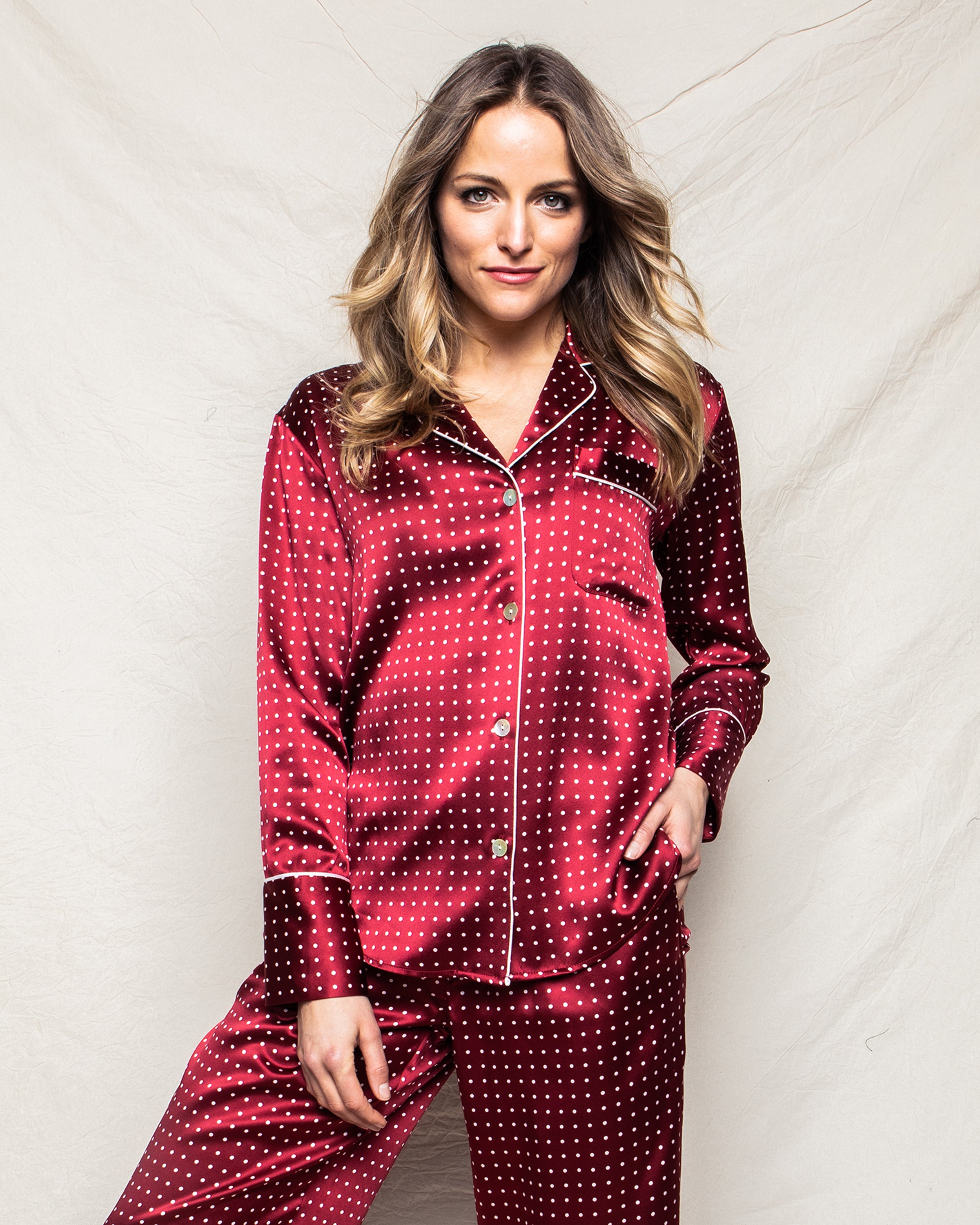 Draped against a light beige backdrop, a person with long hair wears the luxurious Womens Silk Pajama Set in Bordeaux Polka Dot by Petite Plume, with one hand in their pocket and the other arm relaxed by their side.
