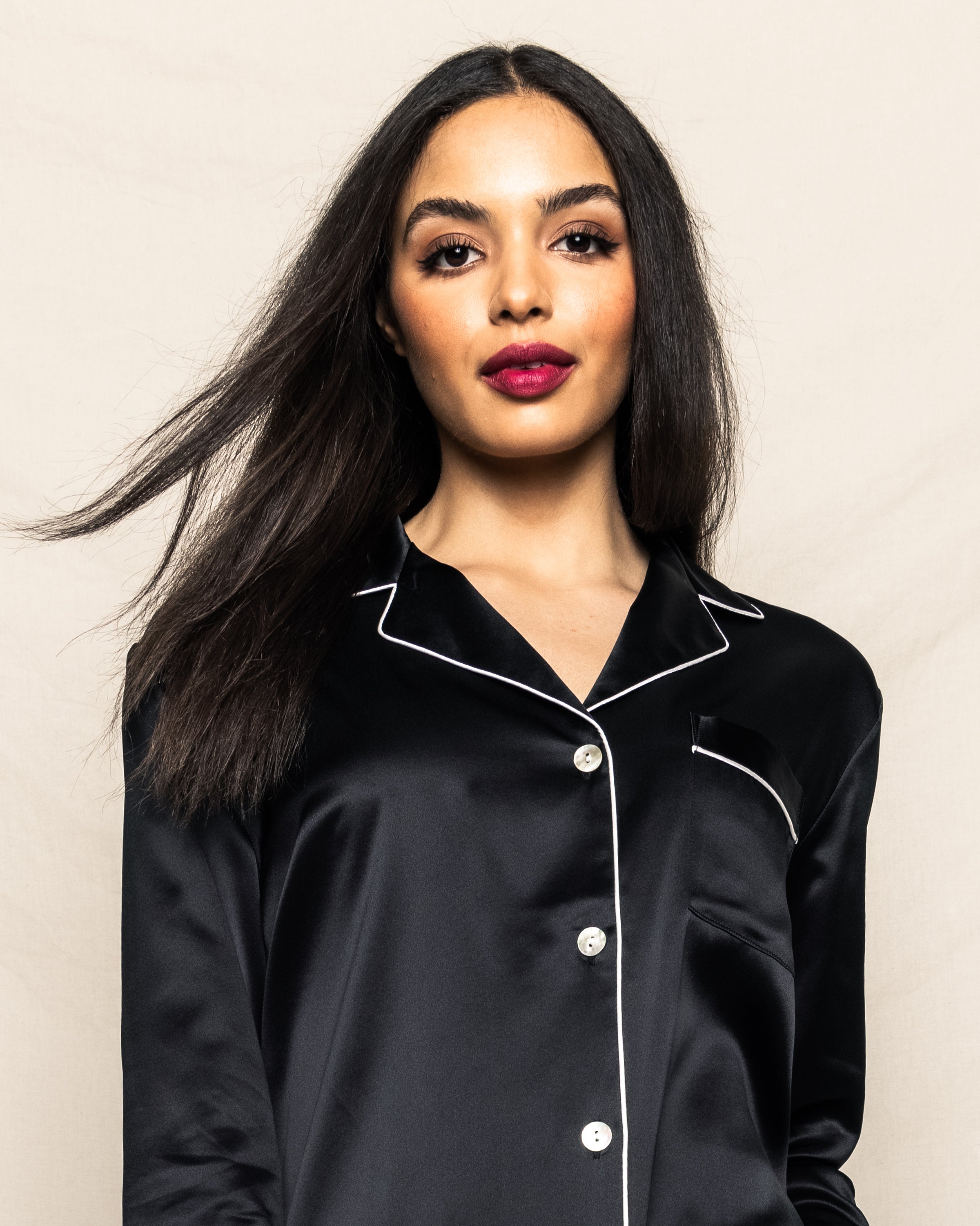 Against a beige backdrop, a woman with long dark hair and bright lipstick exudes confidence in Petite Plumes Womens Silk Pajama Set in Black. The luxurious mulberry silk shirt features white piping, enhancing her graceful look as her hair gently flows.