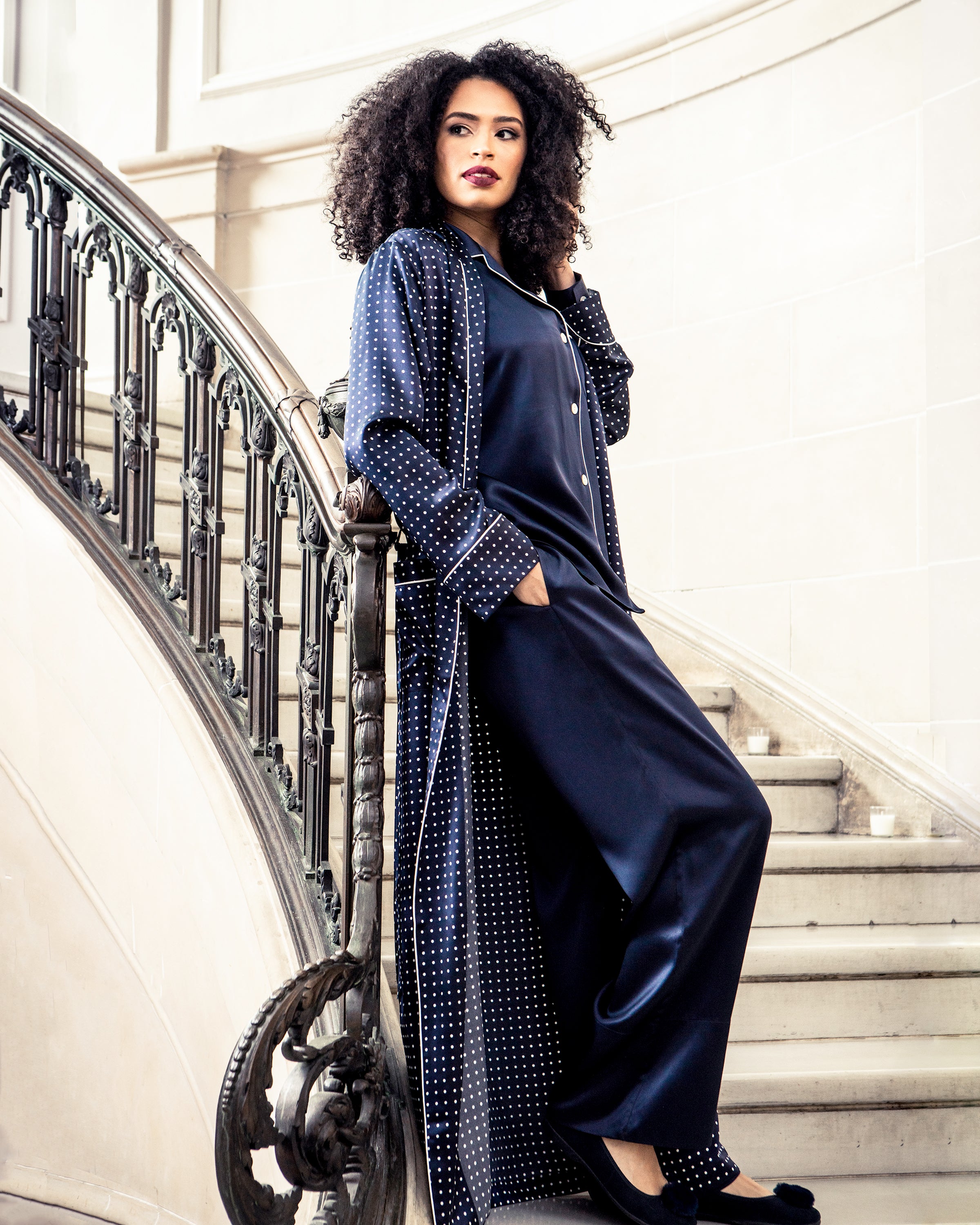 A person with curly hair stands on a staircase in Petite Plumes Womens Silk Pajama Set in Navy, the timeless silhouette complementing the ornate railing and light-colored walls, creating an elegant setting reminiscent of luxury sleepwear.