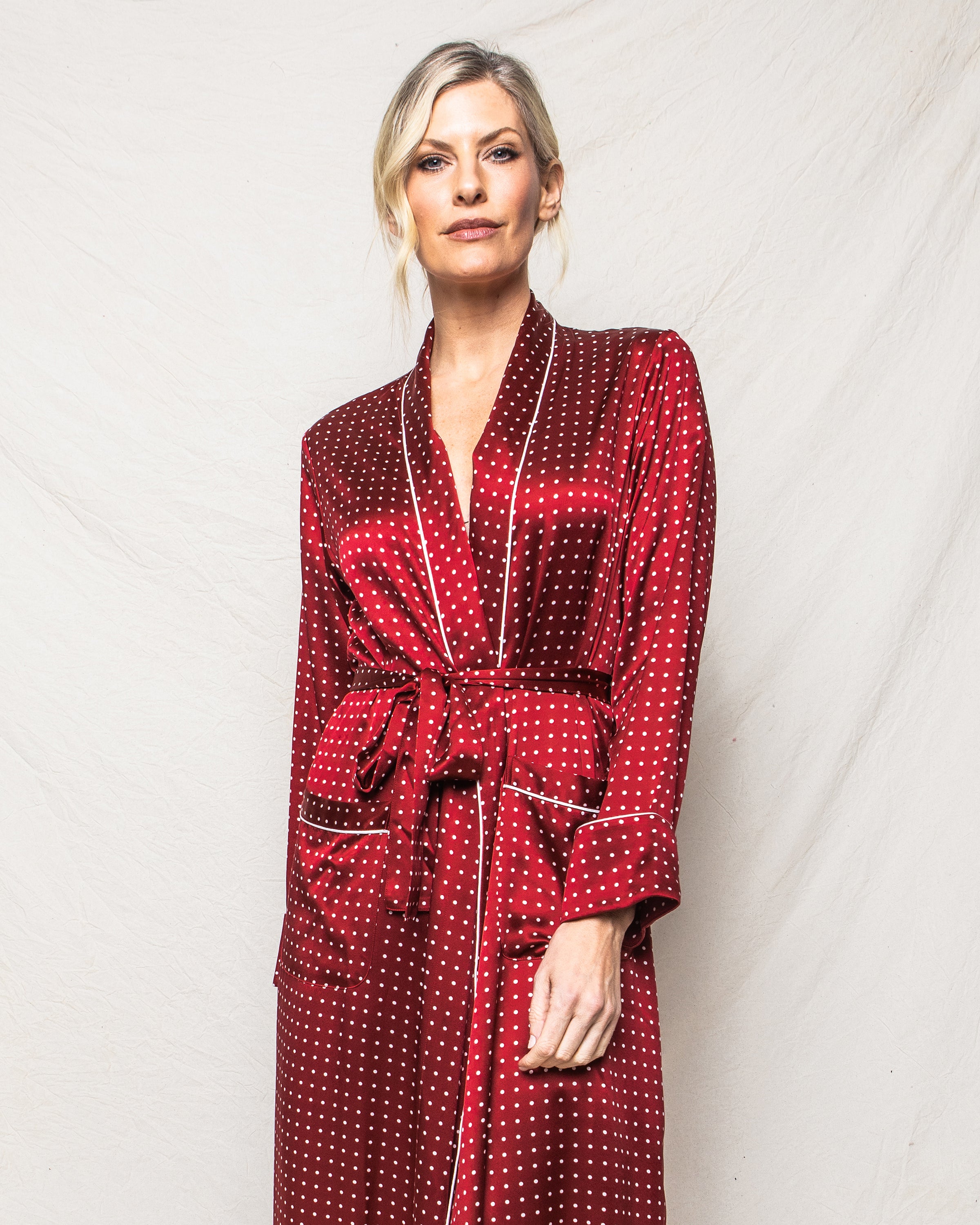 Against a light background, a person with short blonde hair and a relaxed expression wears the luxurious Petite Plume Womens Silk Long Robe in Bordeaux Polka Dot, complete with a matching belt.