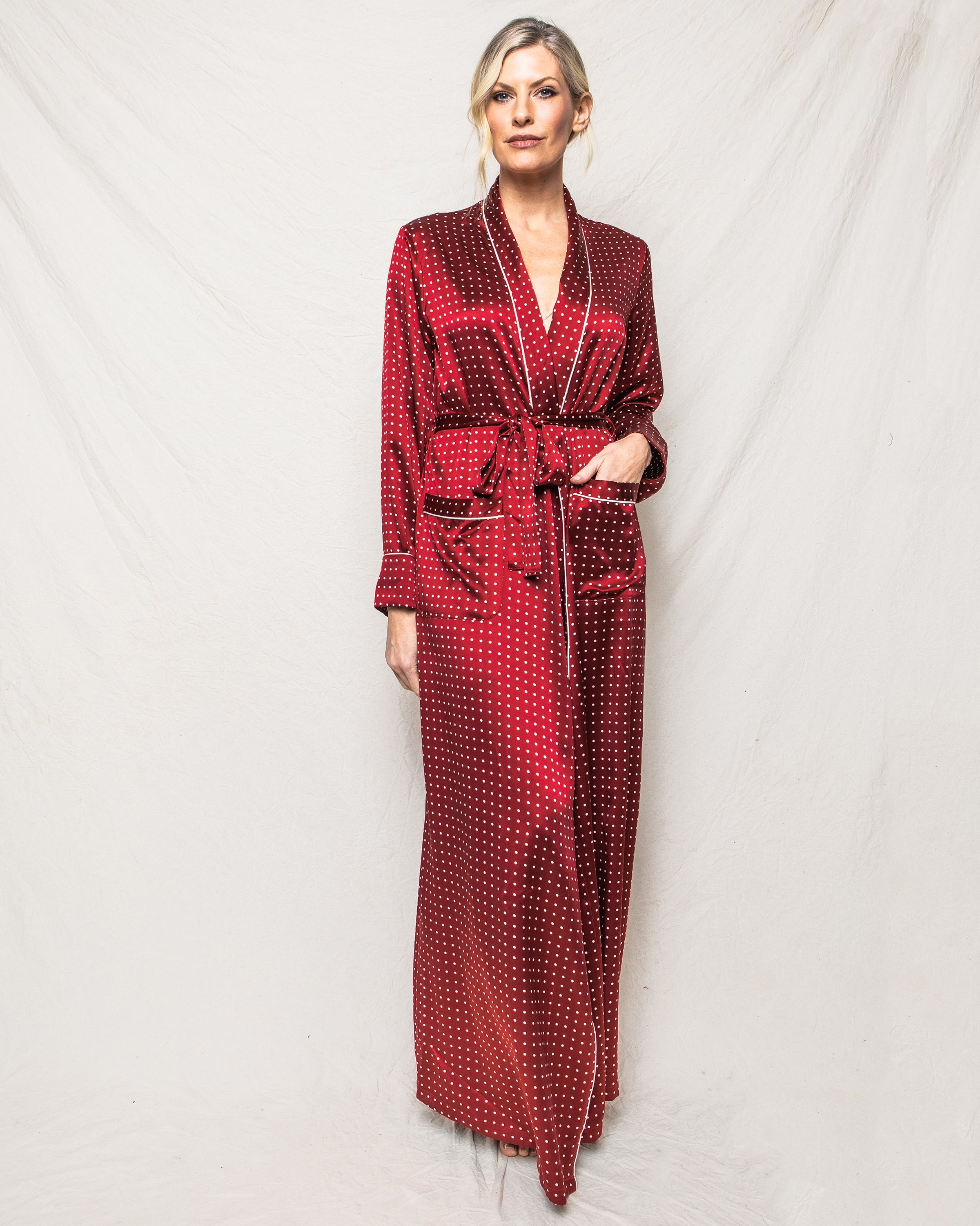 Against a light background, someone stands exuding elegance in the Womens Silk Long Robe in Bordeaux Polka Dot by Petite Plume. Featuring a belted waist and two pockets, their blonde hair is styled up as they gaze towards the camera in this luxurious sleepwear.