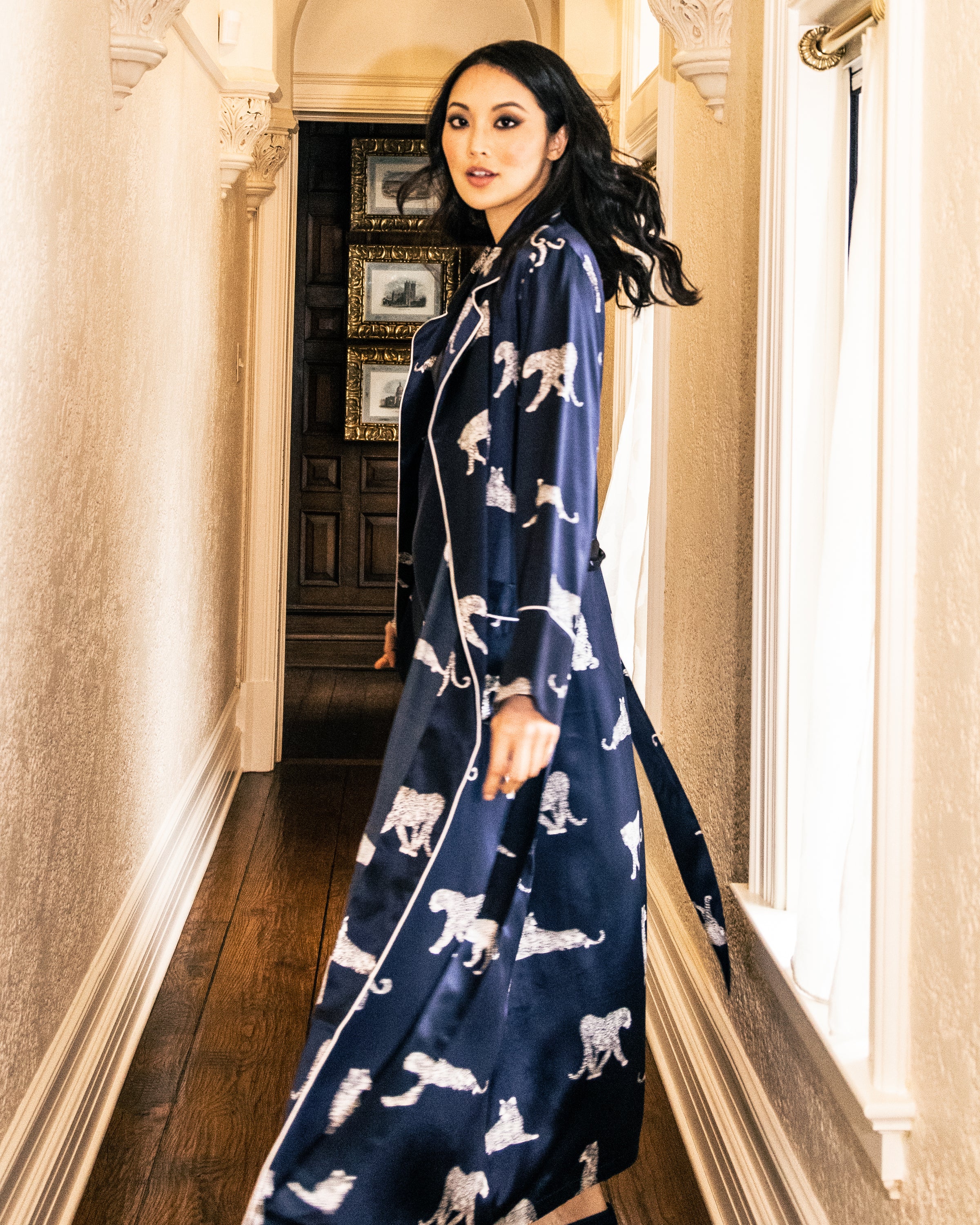 A woman in a luxurious Petite Plume Womens Silk Long Robe in Panthère de Nuit strolls down a narrow wooden-floored hallway. Her hair, styled in classic Hollywood waves, flows elegantly amid the framed pictures on the walls.