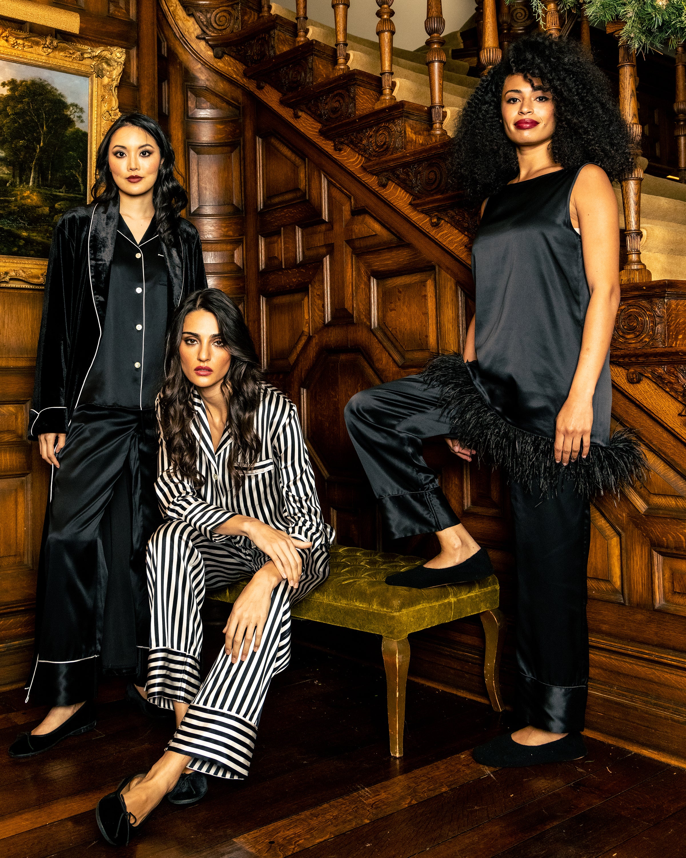 Three women pose on a wooden staircase in luxury sleepwear. One wears Petite Plumes Womens Silk Pajama Set in Black, another sits in striped pajamas, and the third stands in a satin outfit with feather accents. The elegant, vintage setting complements their exquisite ensembles.