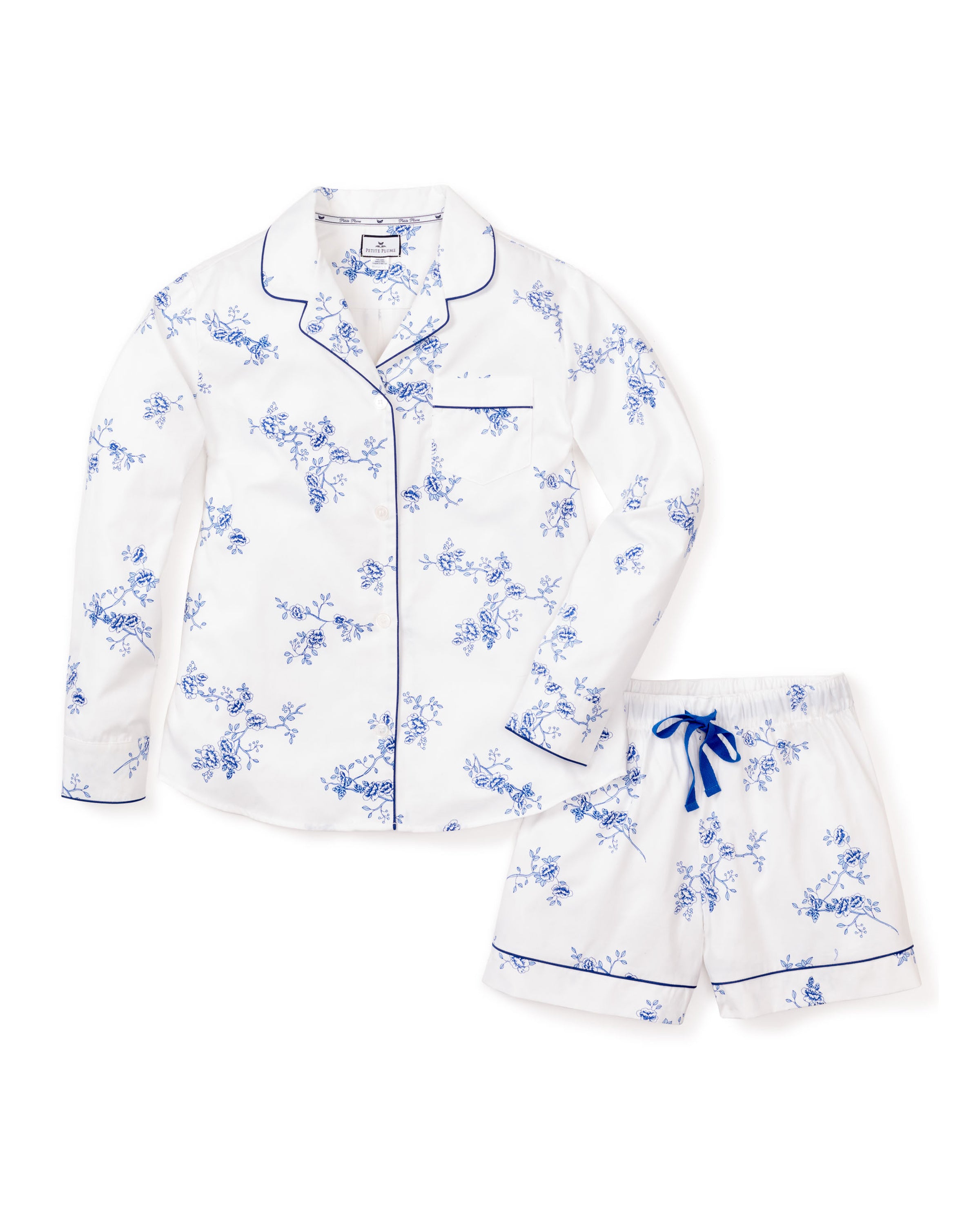 The Womens Twill Long Sleeve Short Set in Indigo Floral by Petite Plume features white PJs with blue floral prints, a long-sleeved button-up top with a pocket and notched collar, and matching shorts with a blue drawstring and piping on the hems. Crafted from premium cotton for luxurious comfort.