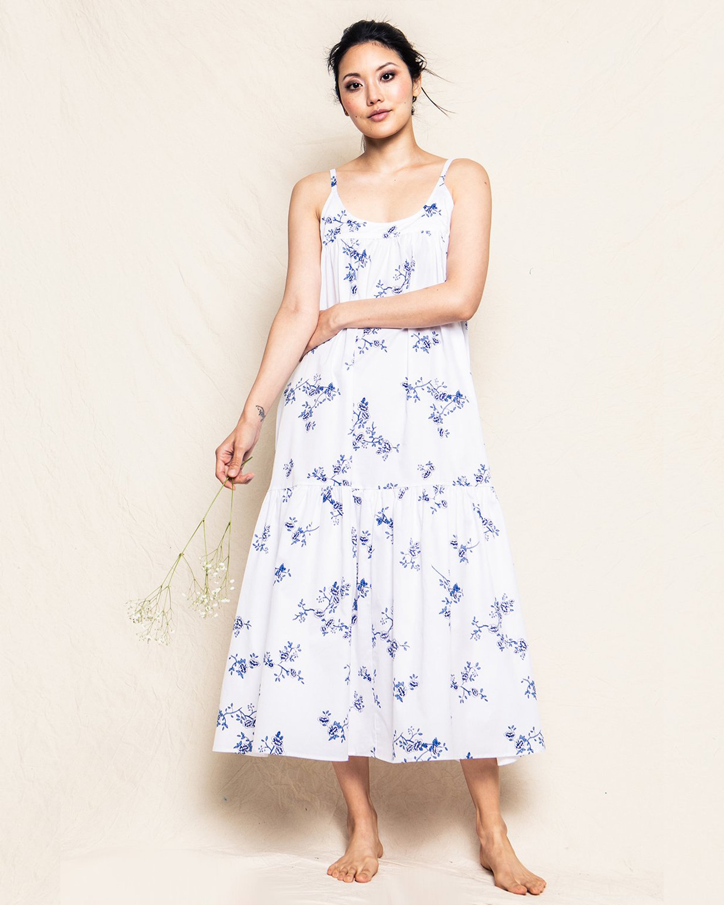 Against a cream background, a barefoot woman dons the Petite Plume Womens Twill Chloé Nightgown in Indigo Floral, featuring blue floral patterns. Her dark hair is elegantly styled up as she holds delicate white flowers.