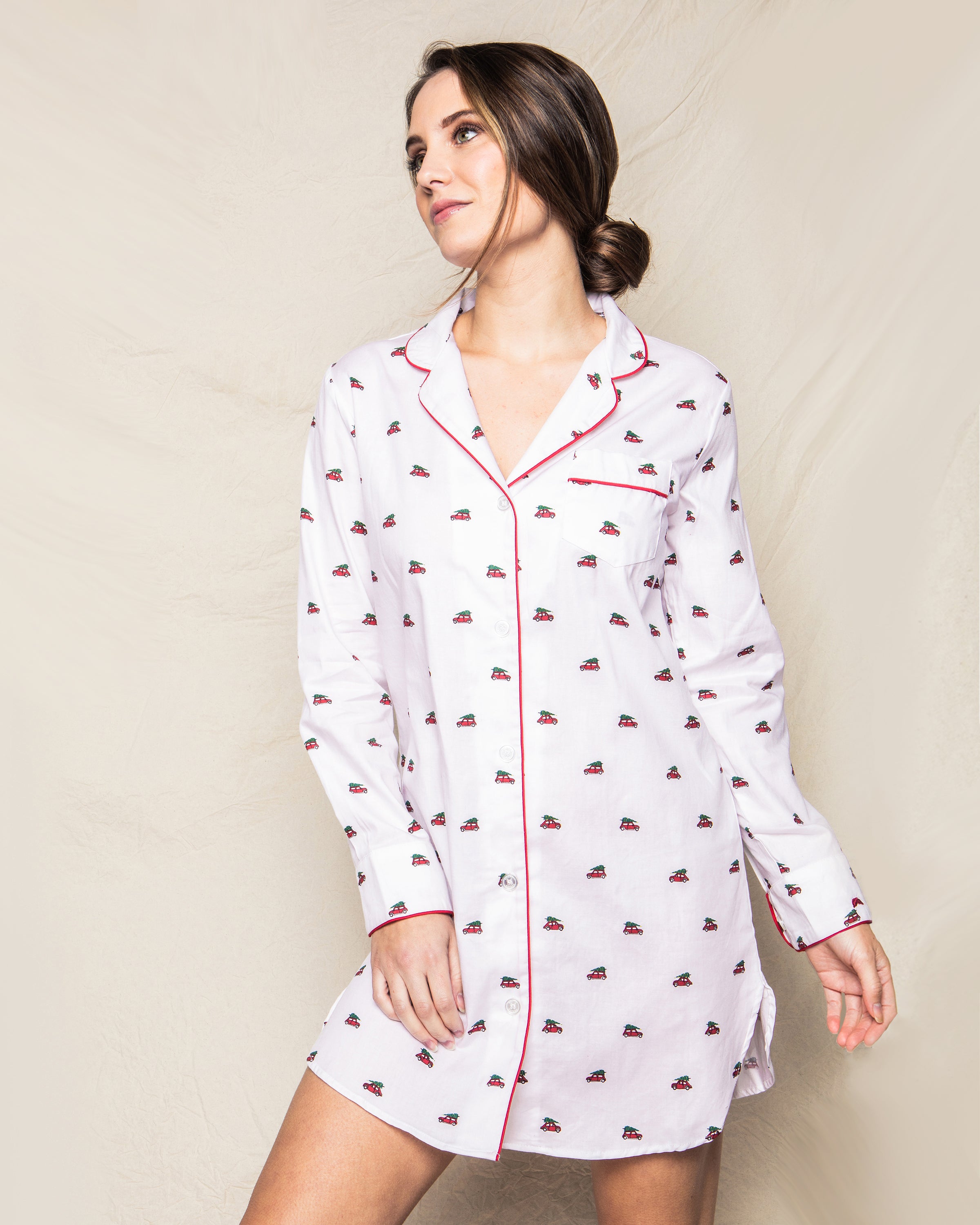 A woman in Petite Plumes Womens Twill Nightshirt in Holiday Journey, featuring a white pattern and red trim, poses against a plain background. Her hair is styled in a loose bun as she looks to the side. The 100% cotton twill fabric ensures comfort, ideal for relaxation.