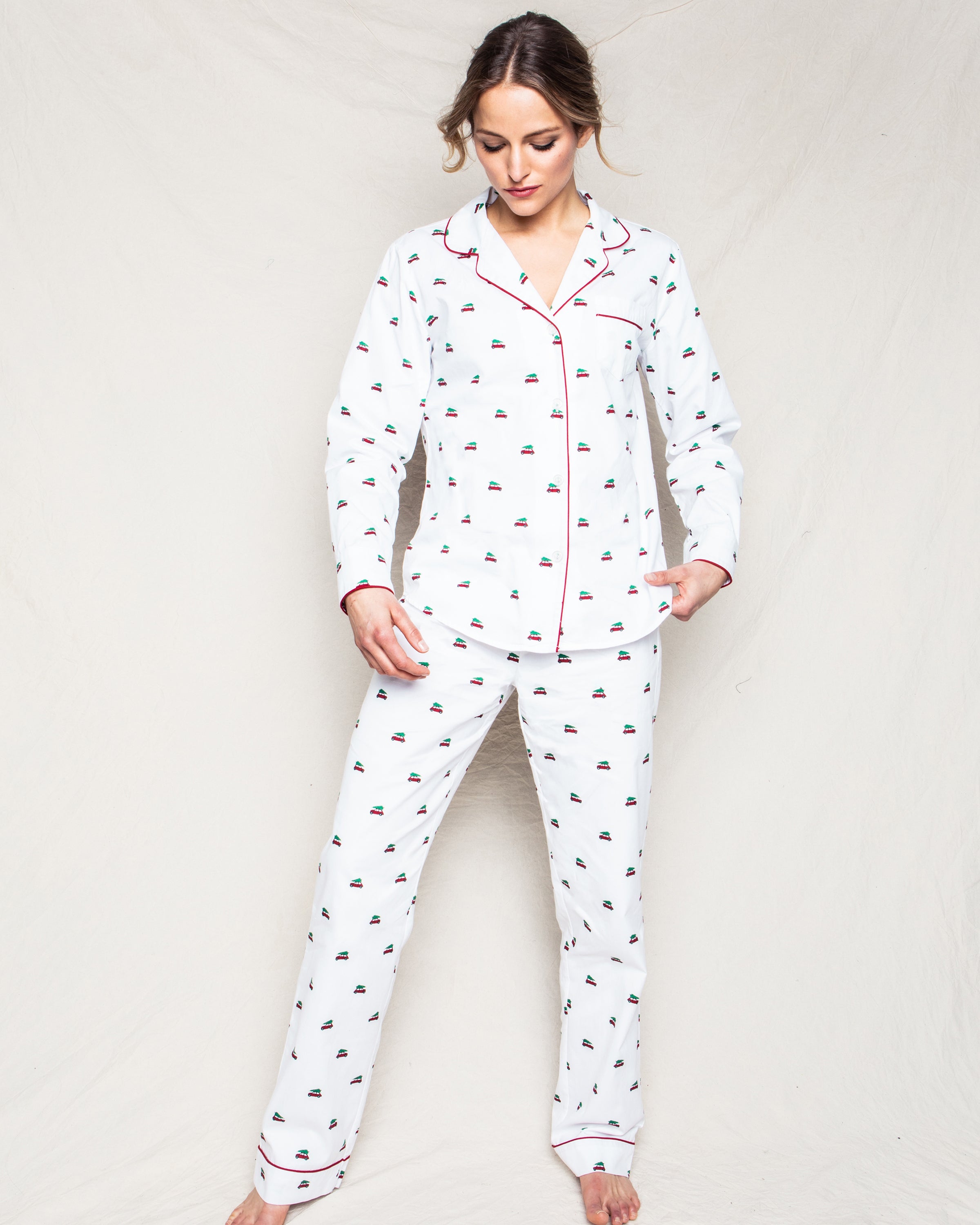 A woman in Petite Plumes Womens Twill Pajama Set in Holiday Journey, featuring a small pattern and red trim, stands against a plain backdrop. She looks down with a relaxed expression, hands in pockets, and hair tied back, exuding a cozy vibe.