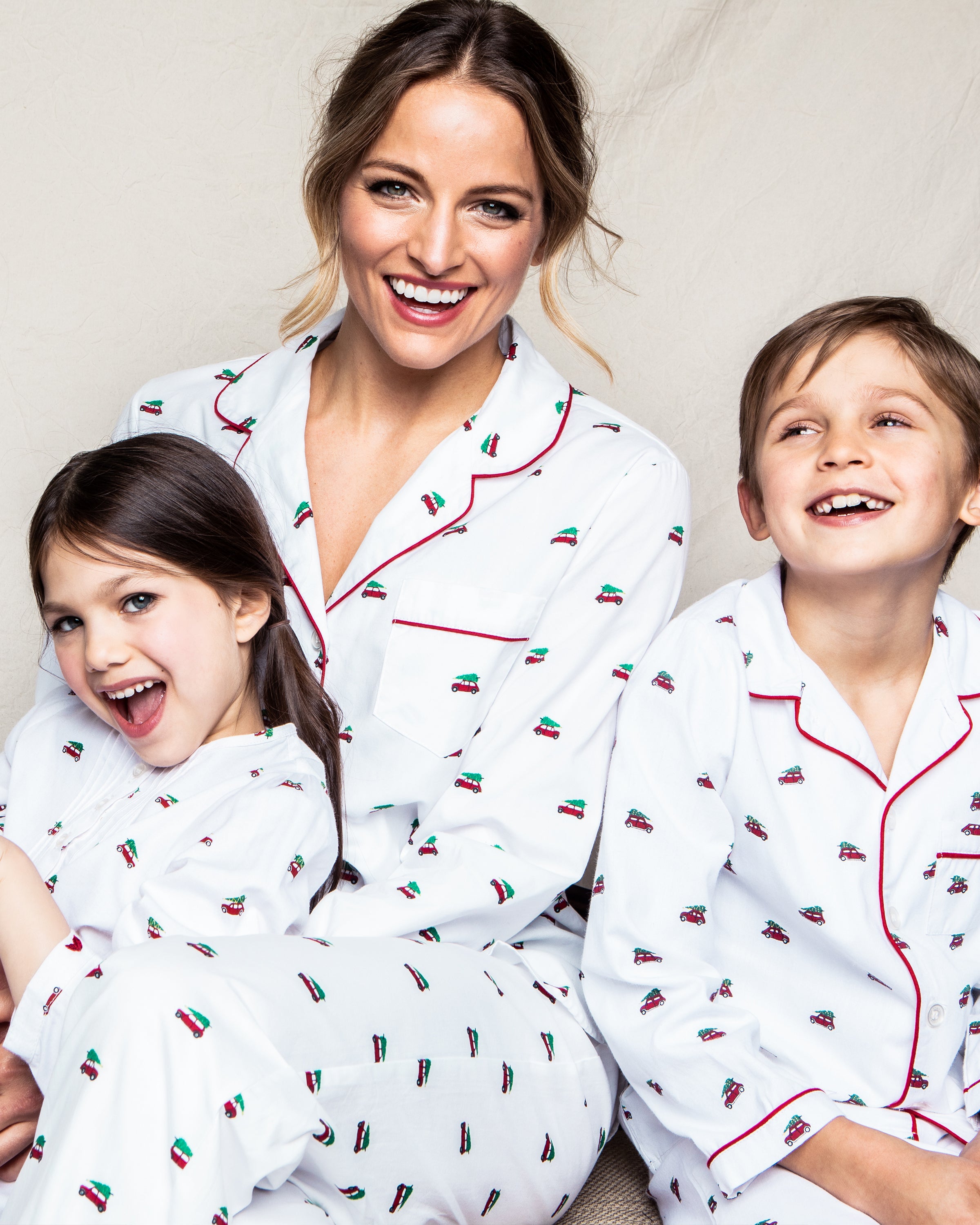 A woman and two children joyfully smile in Petite Plumes Womens Twill Pajama Set in Holiday Journey with matching white cotton, small red cars, red trim, against a beige backdrop, capturing the essence of a delightful holiday.