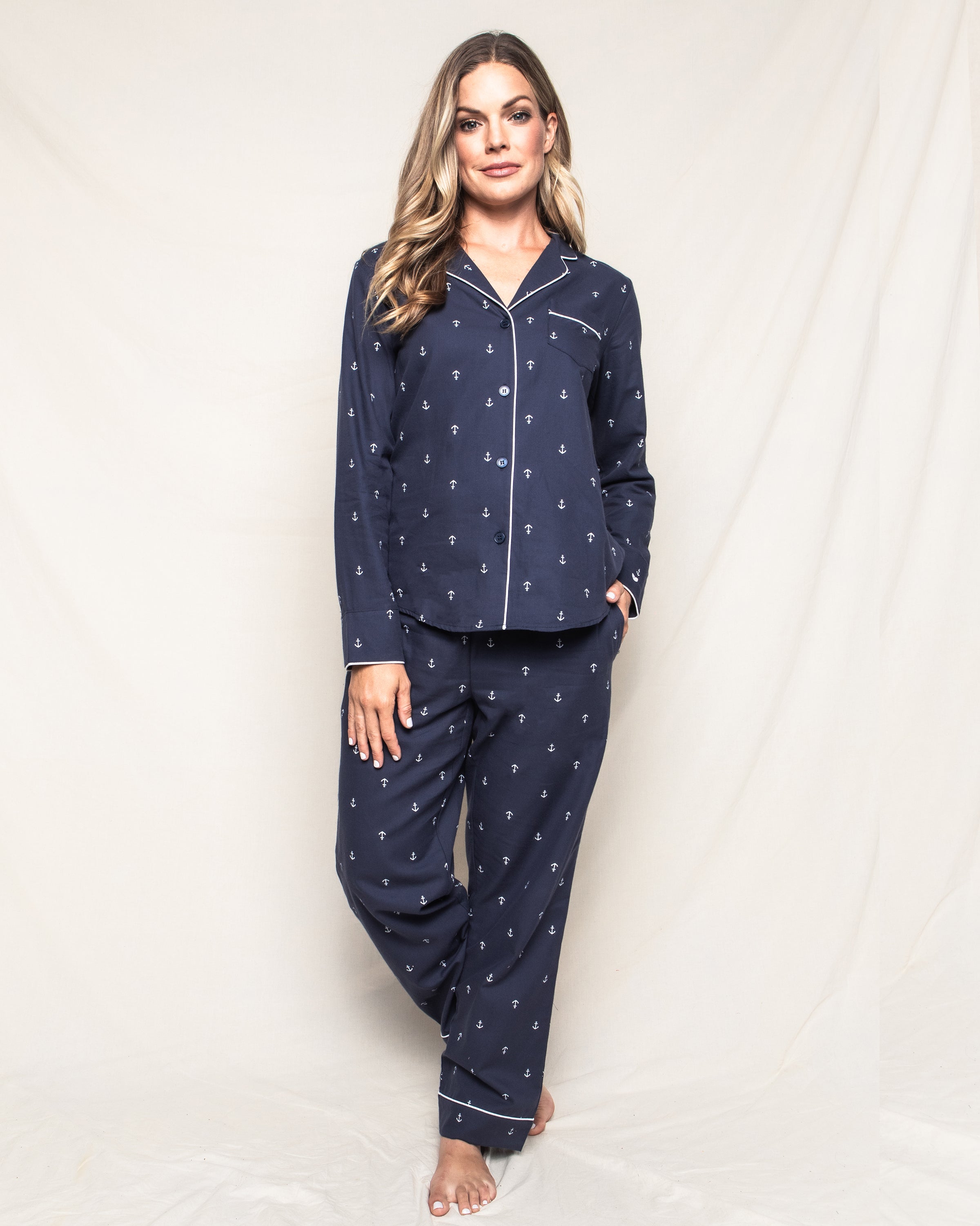 Against a neutral background, an individual models the Petite Plume Womens Twill Pajama Set in Portsmouth Anchors, featuring a navy anchors pattern on fine cotton with a button-up top and matching pants. They have long hair and gaze at the camera with one hand in their pocket.