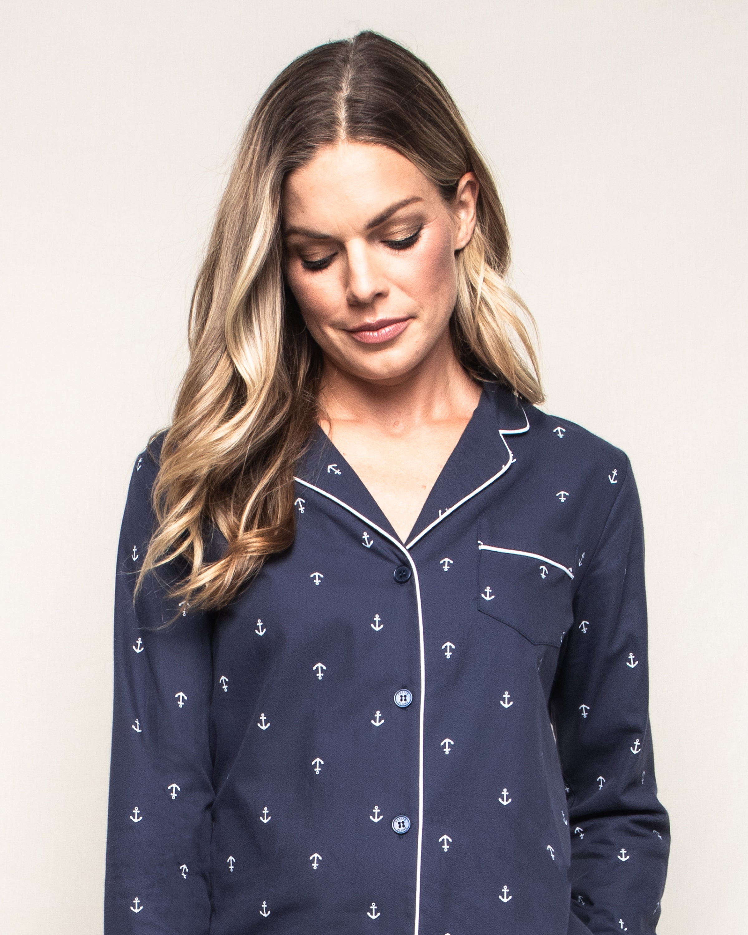 A woman with long, wavy blonde hair wears the Womens Twill Pajama Set in Portsmouth Anchors by Petite Plume, featuring a navy blue top patterned with intricate navy anchors. Against a plain background, she gazes down gently, exuding comfort and grace in this quality cotton ensemble.
