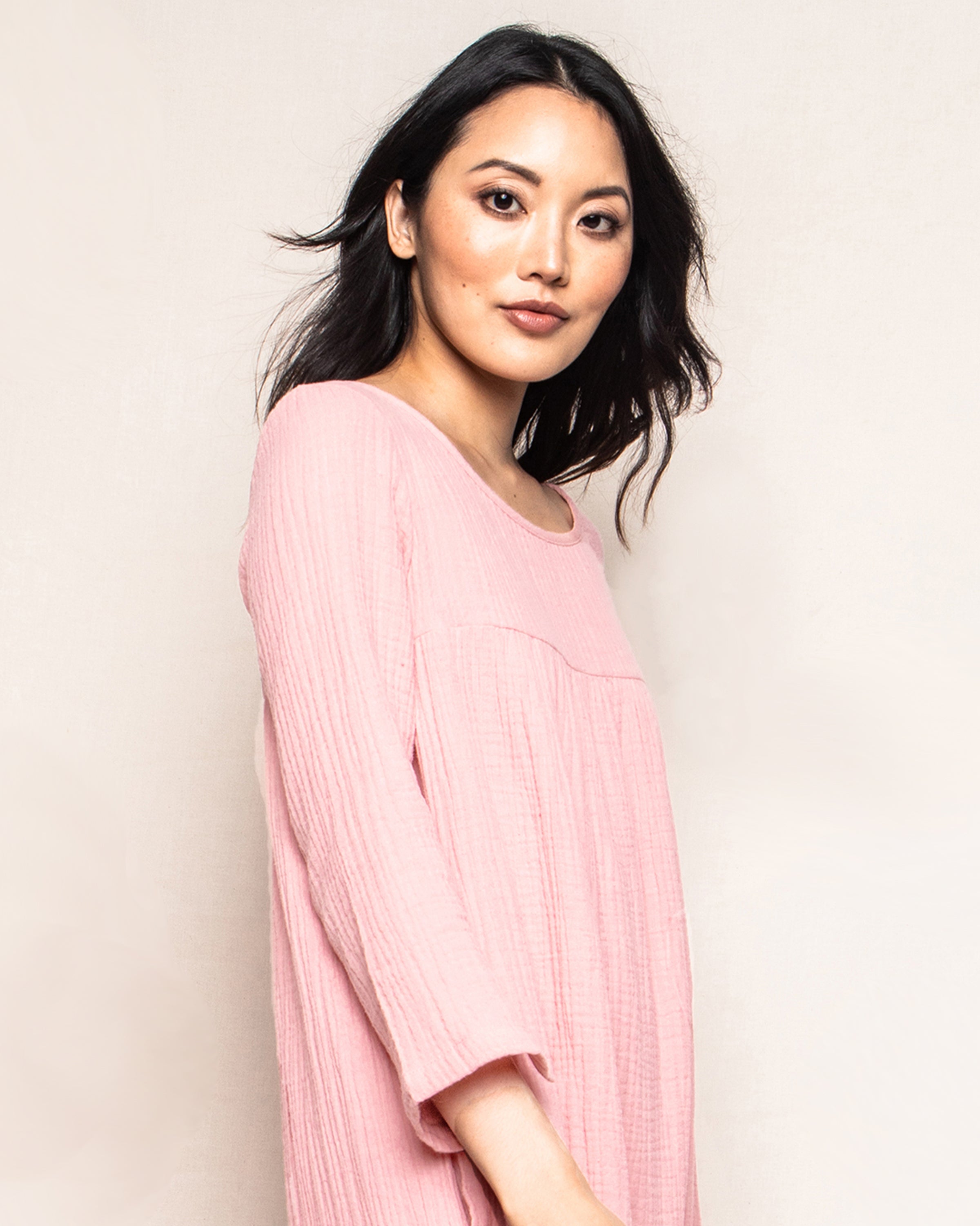 A person with long, wavy dark hair stands against a plain background, dressed in a light pink Womens Gauze Provence Sundress by Petite Plume. Their confident expression and side gaze capture the essence of luxury loungewear.