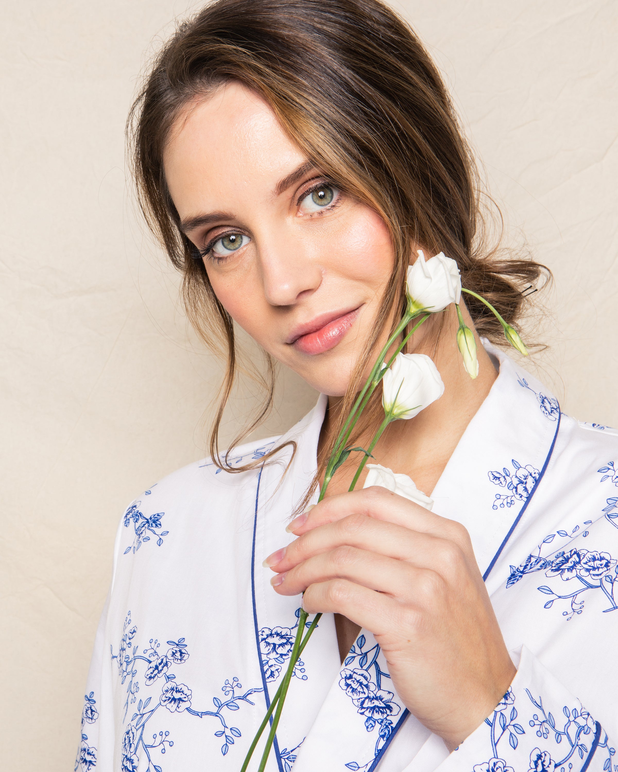 A woman with light brown hair gently holds white flowers near her face, wearing a Petite Plume Womens Twill Robe in Indigo Floral. Her expression is gentle against a soft beige background.