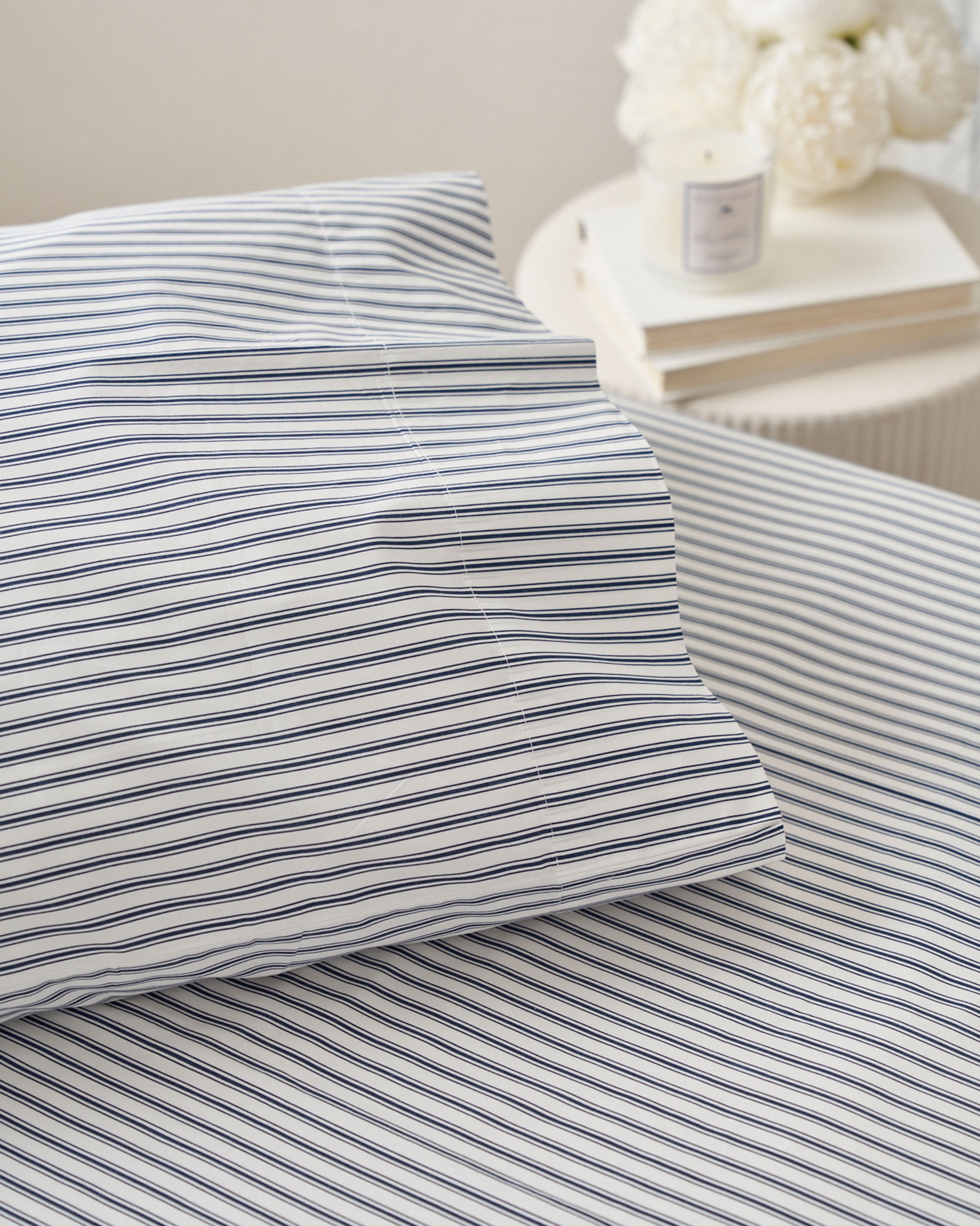 A close-up shows a bed made with Petite Plumes Luxe Premium Cotton Bed Sheets in Navy French Ticking. Nearby, a round white side table holds books, a lit candle, and a flower arrangement, creating a calm ambiance. The 100% cotton fabric provides a luxurious feel.