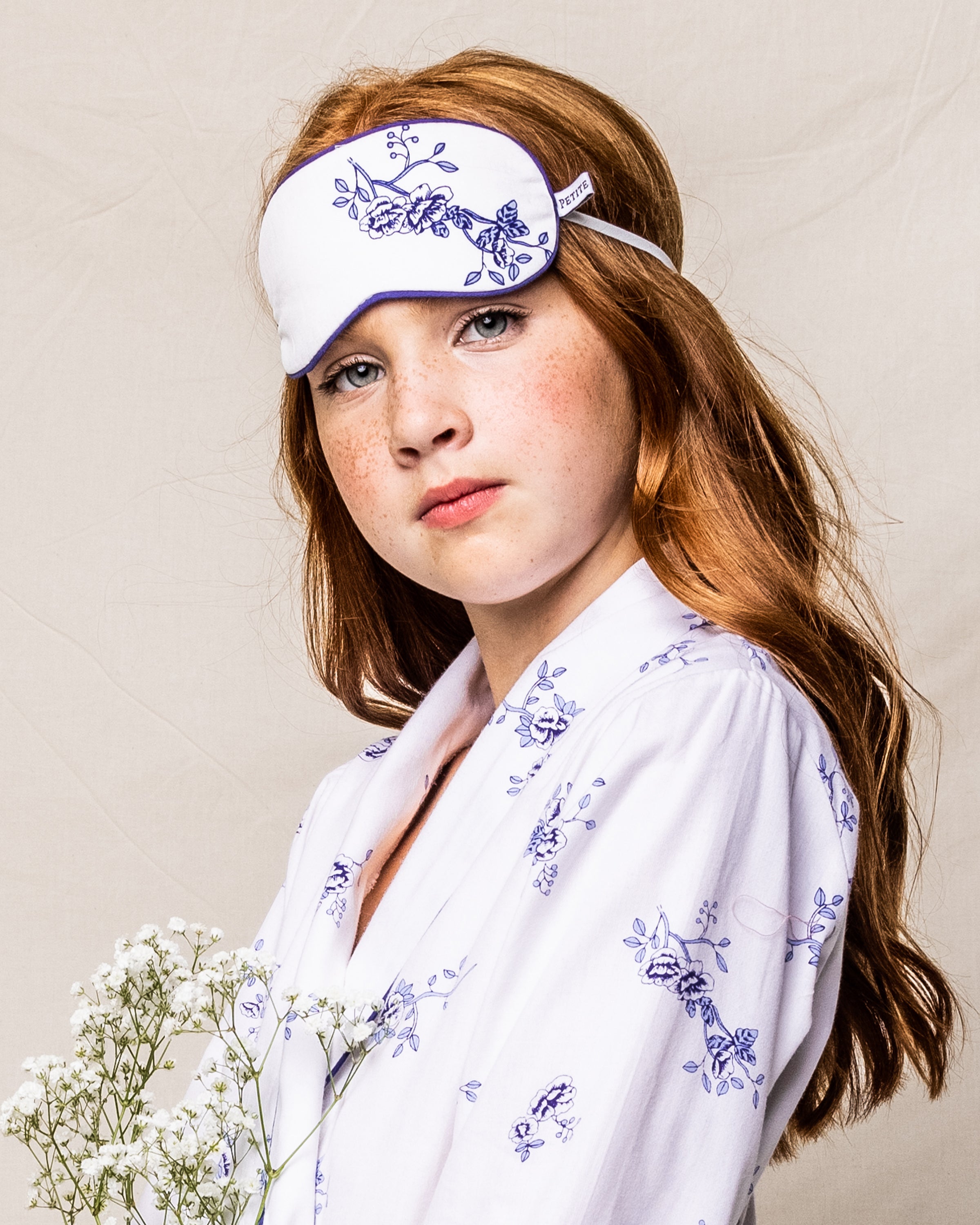 A person with long red hair wears a Kids Sleep Mask in Indigo Floral by Petite Plume, stepping into dreamland. They hold a small bouquet of white flowers against a cream background, styled for elegance.