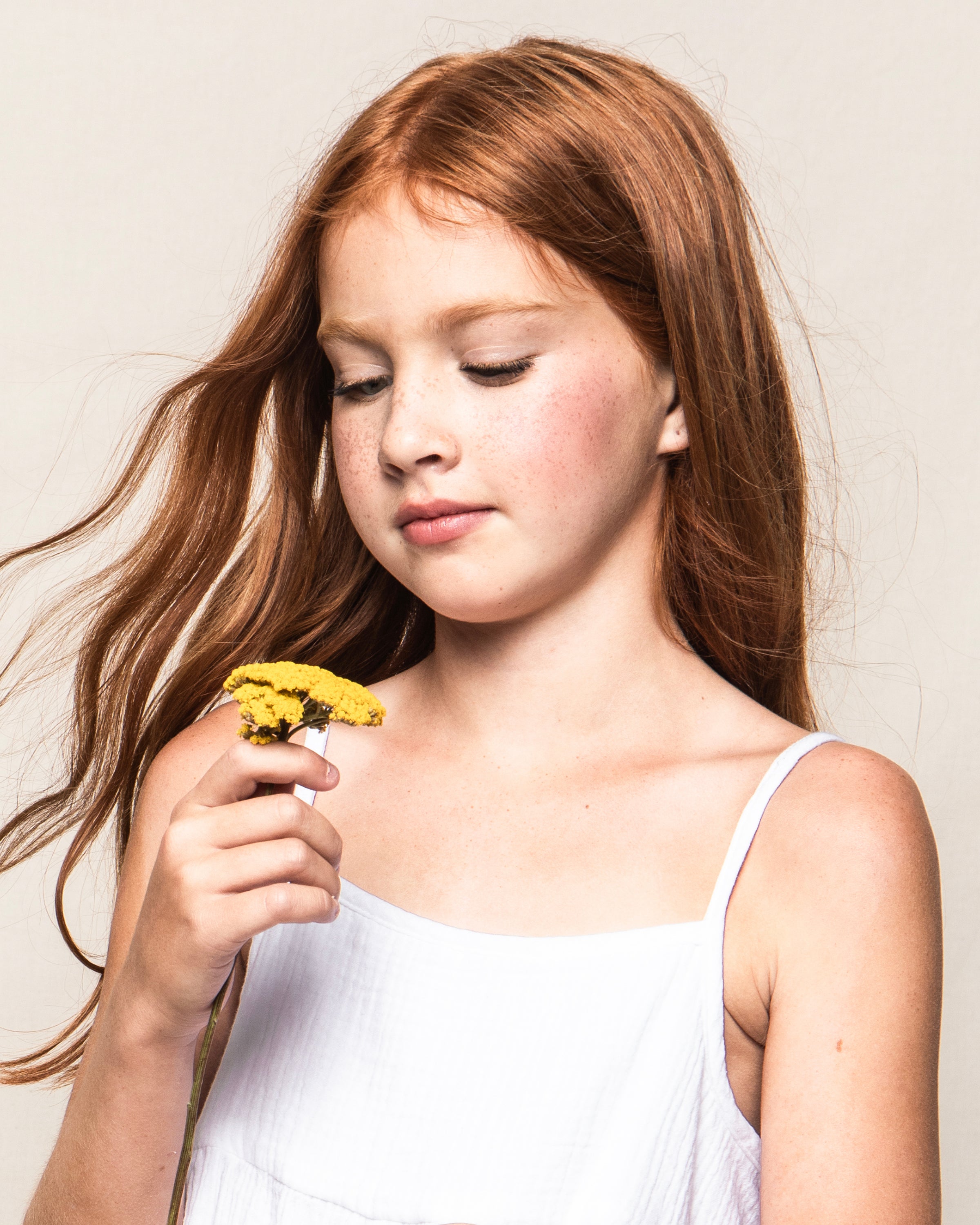 A young girl with long red hair and freckles admires a yellow flower in her hand, wearing Petite Plumes lightweight cotton Girls Gauze Serene Sundress in White, set against a neutral beige background.