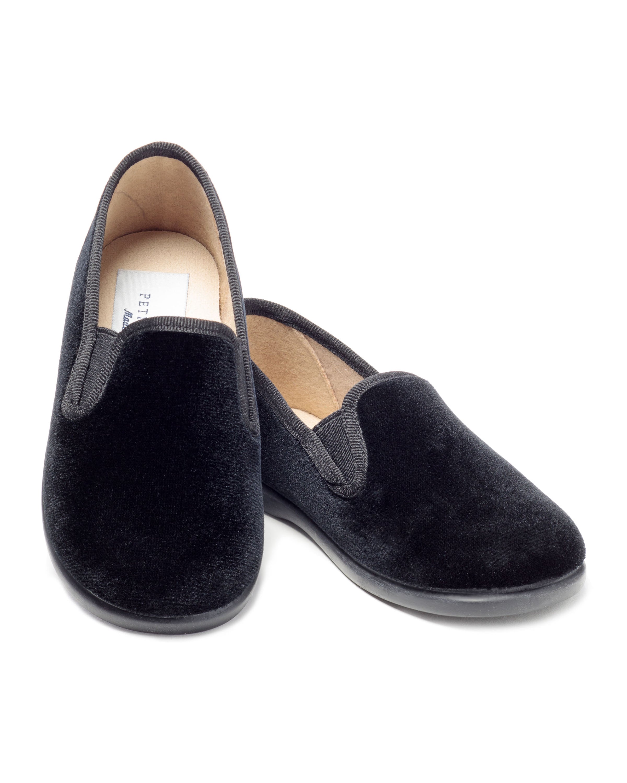 The Kids Gable Slipper by Petite Plume is crafted in black velvet with rounded toes, elastic side inserts, and flat rubber soles. Its cozy beige interior makes these slippers ideal for winter nights, featuring one shoe slightly angled in front of the other.