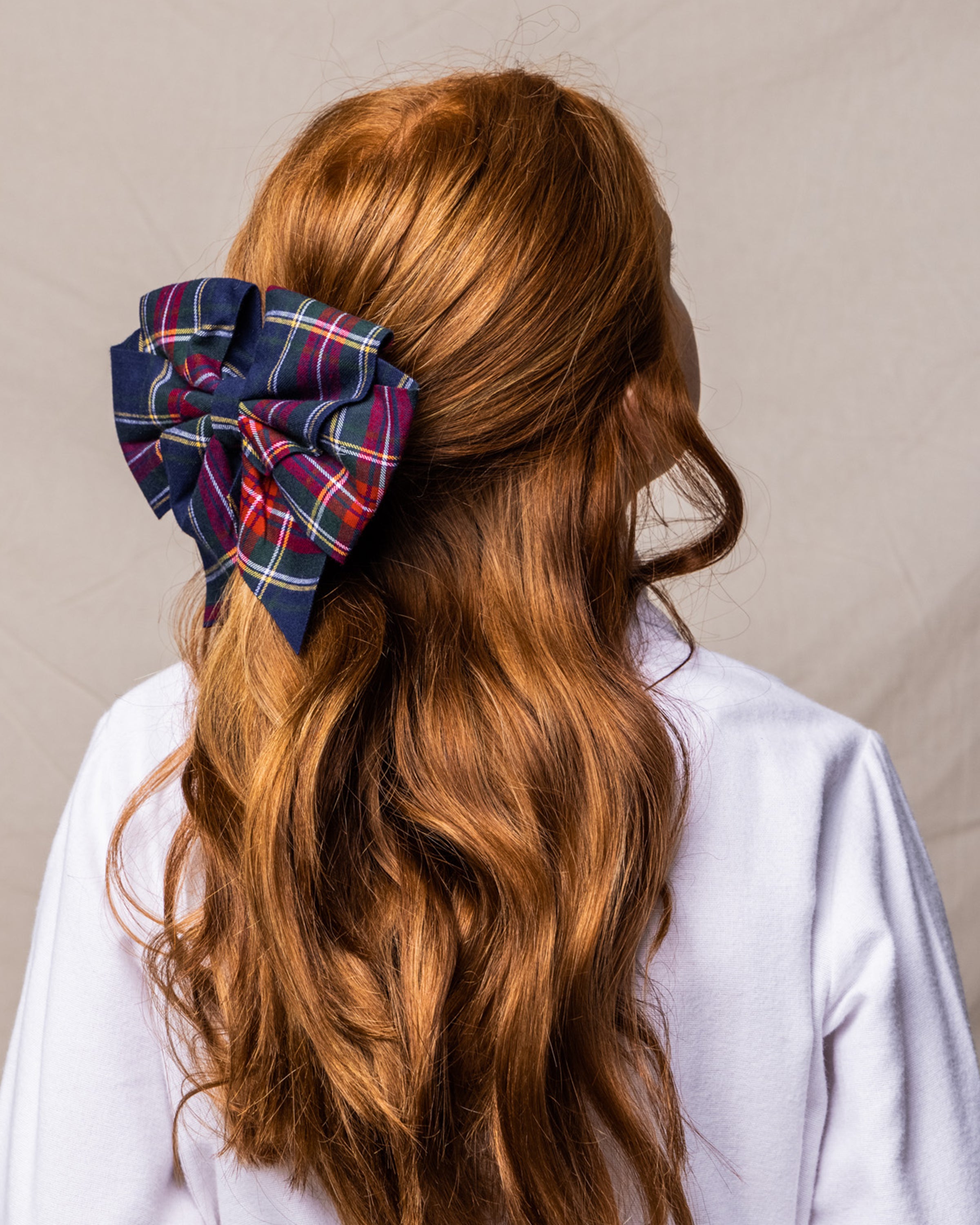 A person with long, wavy red hair, tied with a large Petite Plume Windsor Tartan Girls Hair Bow in navy, red, and white, stands against a neutral backdrop wearing a crisp white shirt—ideal for gifting during the holidays.