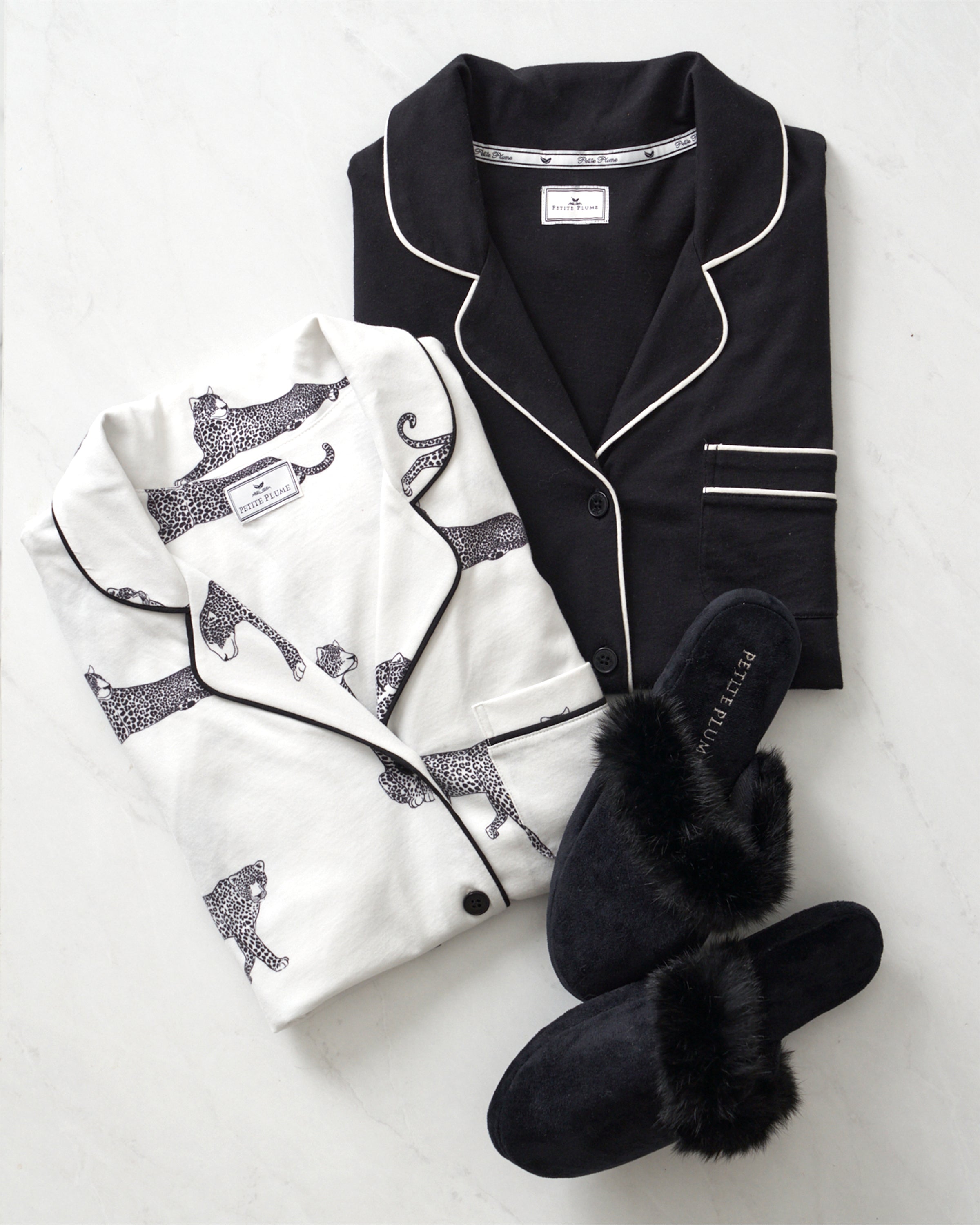 Two folded luxury pajama sets rest on a light surface: the Womens Pima Astaire Pajama Set in Black by Petite Plume and another with black animal prints and piping. Made from Peruvian Pima cotton, they promise comfort. A pair of black fuzzy slippers is in front, completing the cozy ensemble.