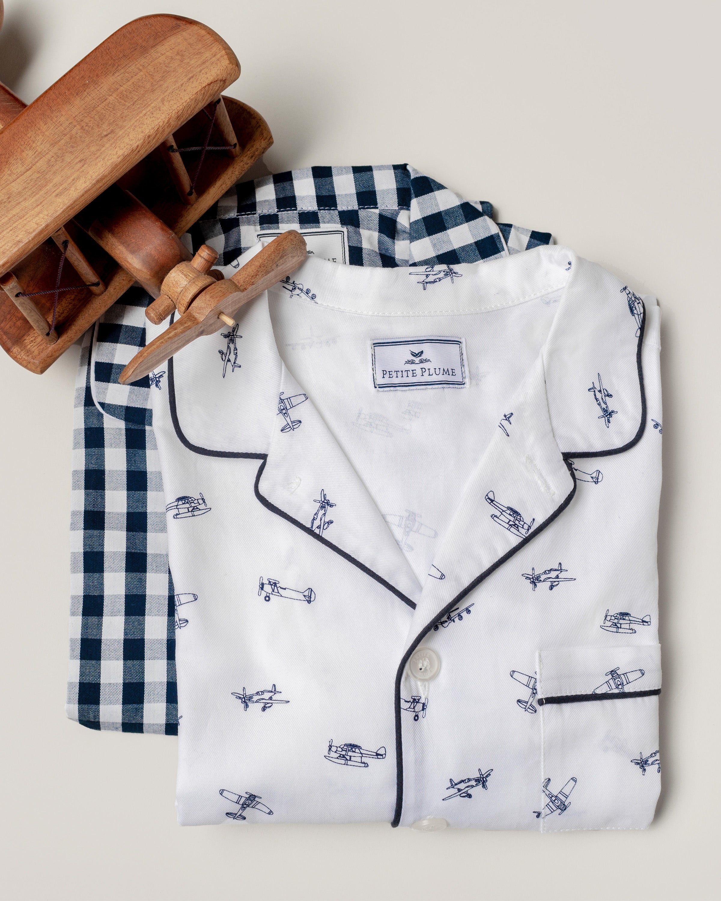 Two folded Petite Plume Kids Twill Pajamas, featuring a Par Avion print with airplanes on top and blue/white checkered bottoms, rest on a neutral background. Beside them lies a wooden toy airplane, perfect for luxurious sleepwear dreams.