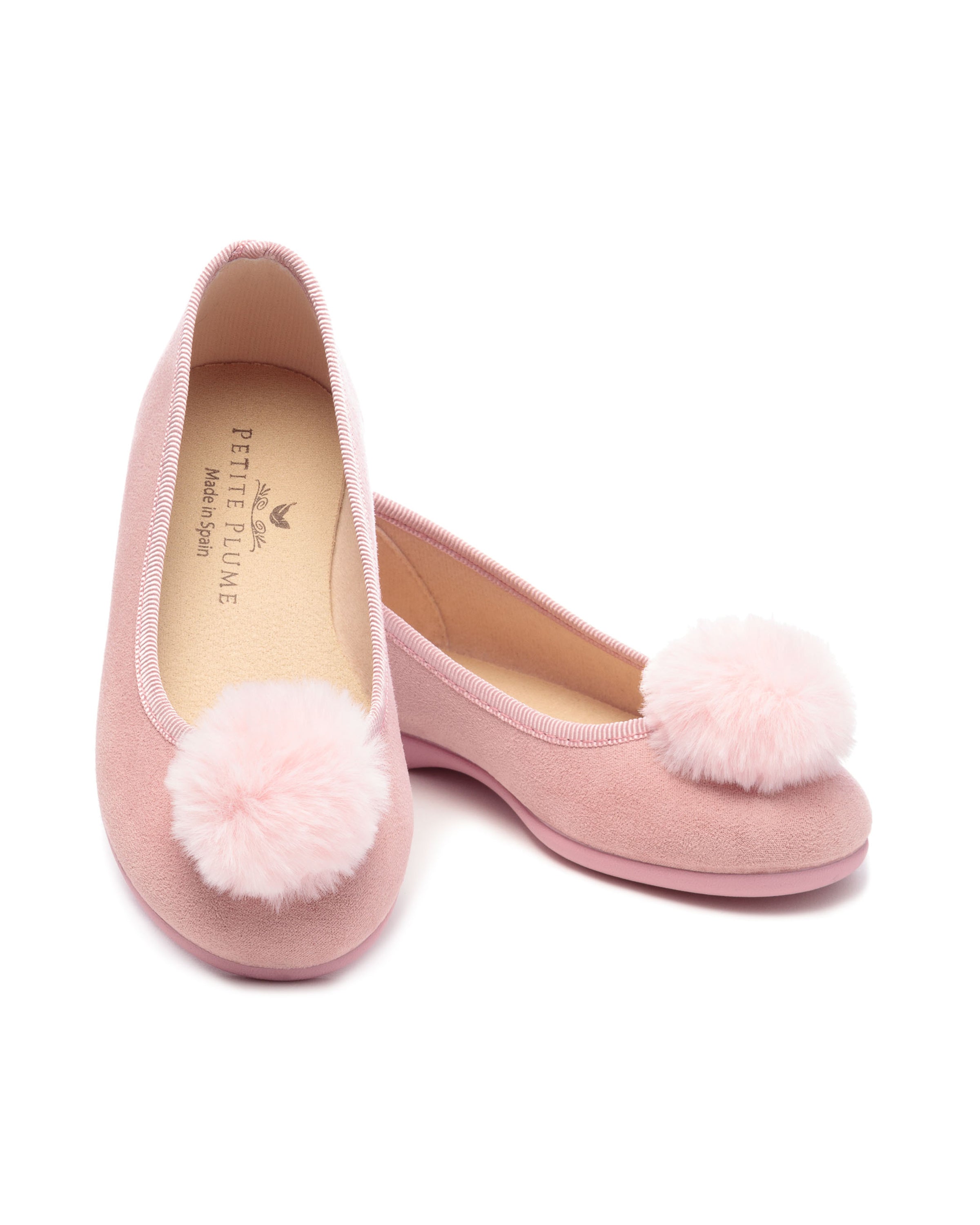 The Kids Juliette Slipper by Petite Plume is made of antique rose suede with festive pom-poms. It features a beige interior with branding, slightly rounded toe, and flat sole.