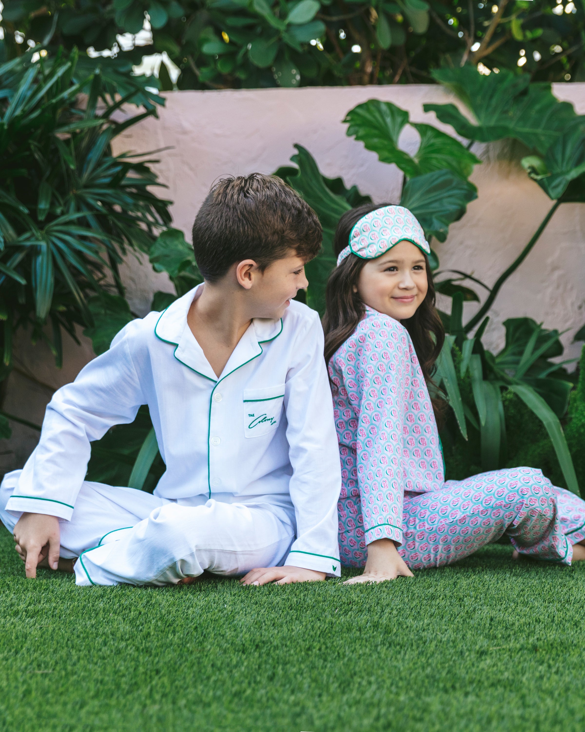 Two kids sit back-to-back on the grass, surrounded by plants. The boy wears a Petite Plume Colony Hotel x Petite Plume Kids White with Green Piping Pajama Set, while the girl sports patterned pajamas and a matching sleep mask, perfectly capturing the essence of luxurious sleepwear.