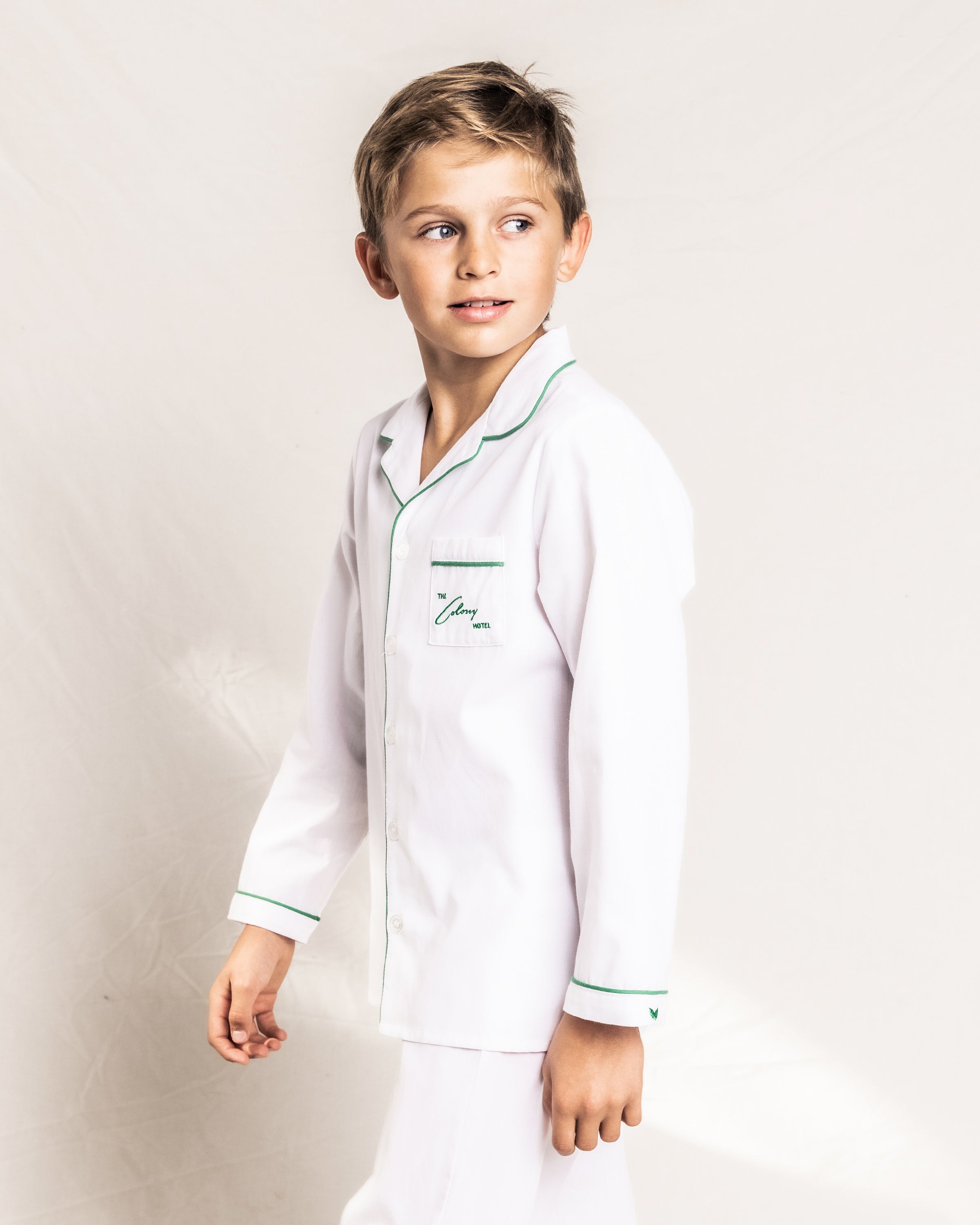 A young boy wears the Colony Hotel x Petite Plume Kids White with Green Piping Pajama Set. His short light brown hair complements his slight side smile as he gazes away, embodying the Colony Hotels classic elegance against a plain white background.
