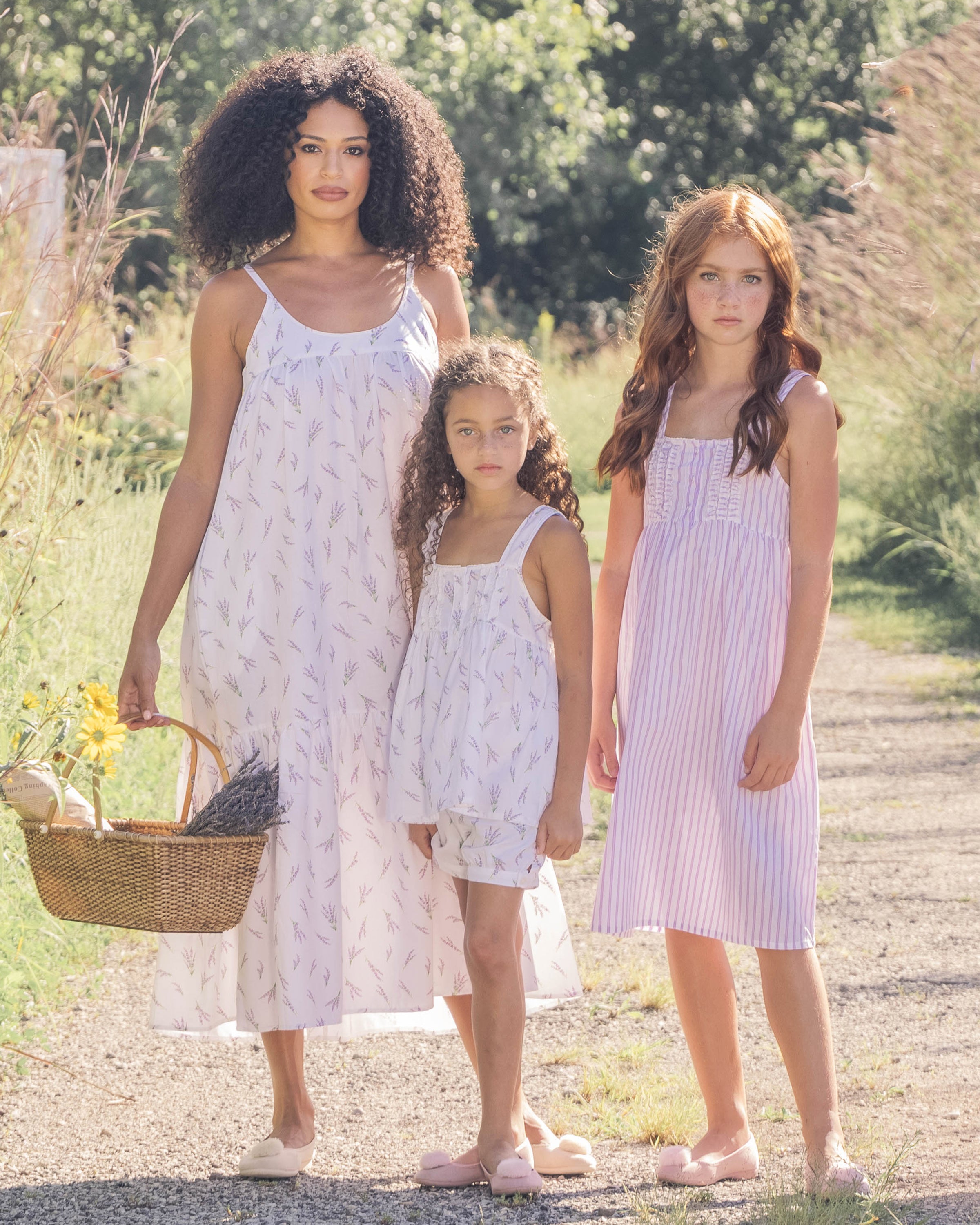 Three girls stand outdoors on a sunny path, surrounded by greenery, wearing matching Petite Plume Girls Twill Charlotte Nightgowns in Lavender French Ticking. The tallest holds a wicker basket filled with yellow flowers. Their curly and wavy hair complements their relaxed, summery look.