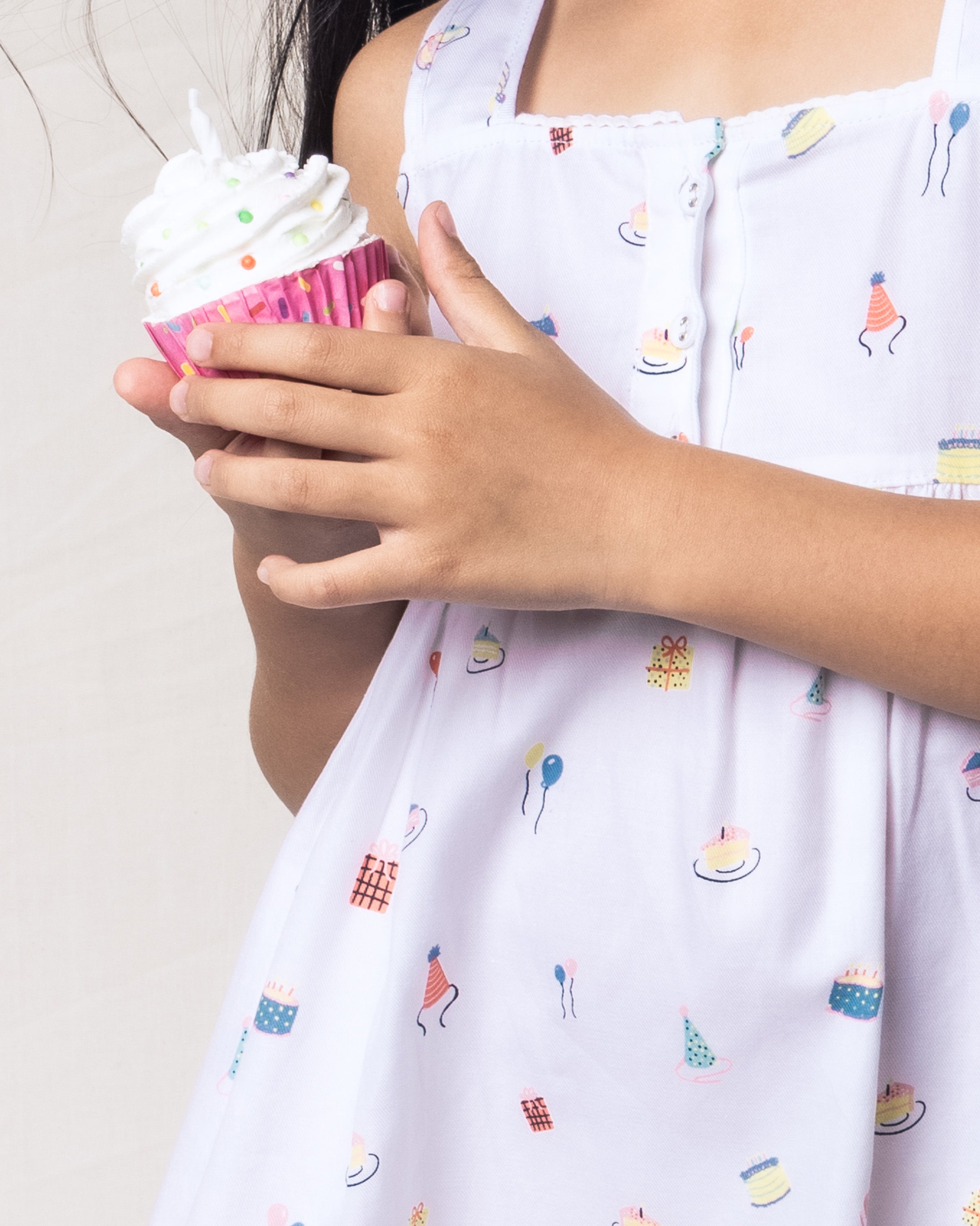 A child in Petite Plumes Girls Twill Charlotte Nightgown in Birthday Wishes, featuring colorful party-themed prints including balloons, cakes, and hats on yarn-dyed cotton, holds a cupcake with white frosting and sprinkles against a neutral background.