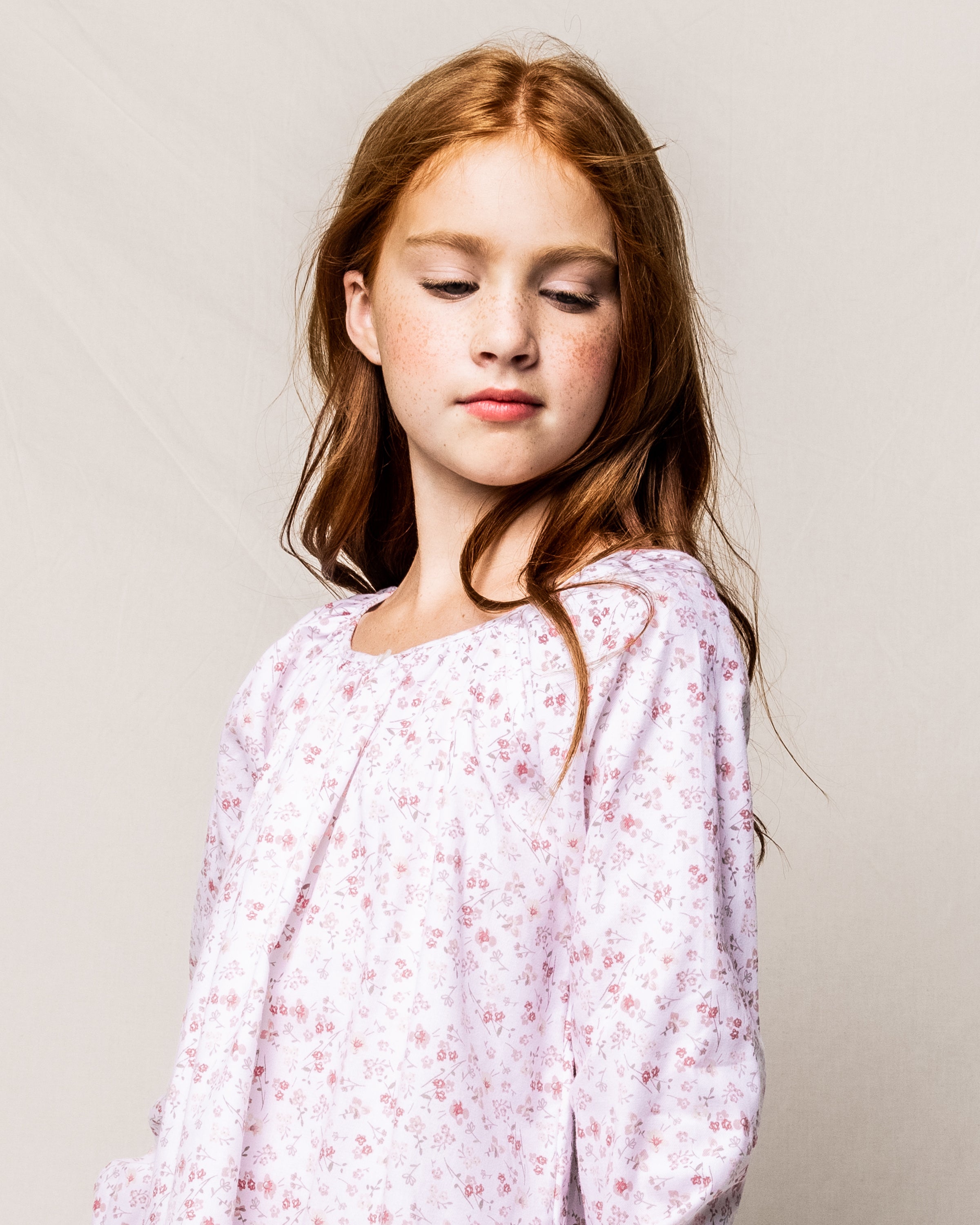 The young girl with long, reddish-brown hair and freckles, dressed in a Girls Twill Delphine Nightgown in Dorset Floral by Petite Plume, gazes downward with a subtle smile. Her white floral long-sleeve top enhances the serene vibe against a neutral background.