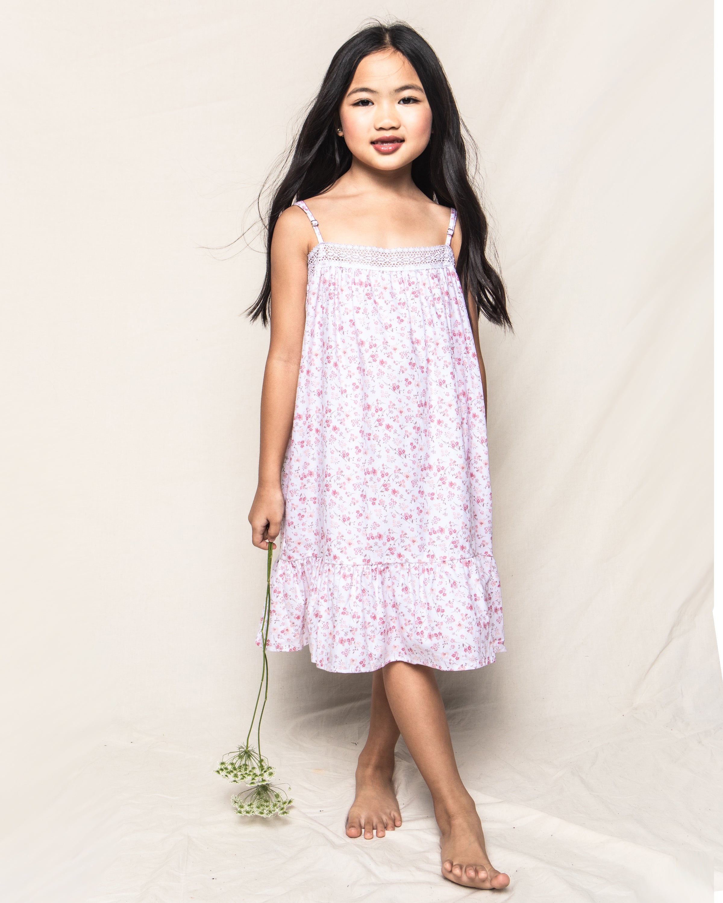 A young girl with long dark hair wears the Dorset Floral design of the Girls Twill Lily Nightgown by Petite Plume. She holds a small bouquet and stands barefoot on a light fabric backdrop, smiling softly.