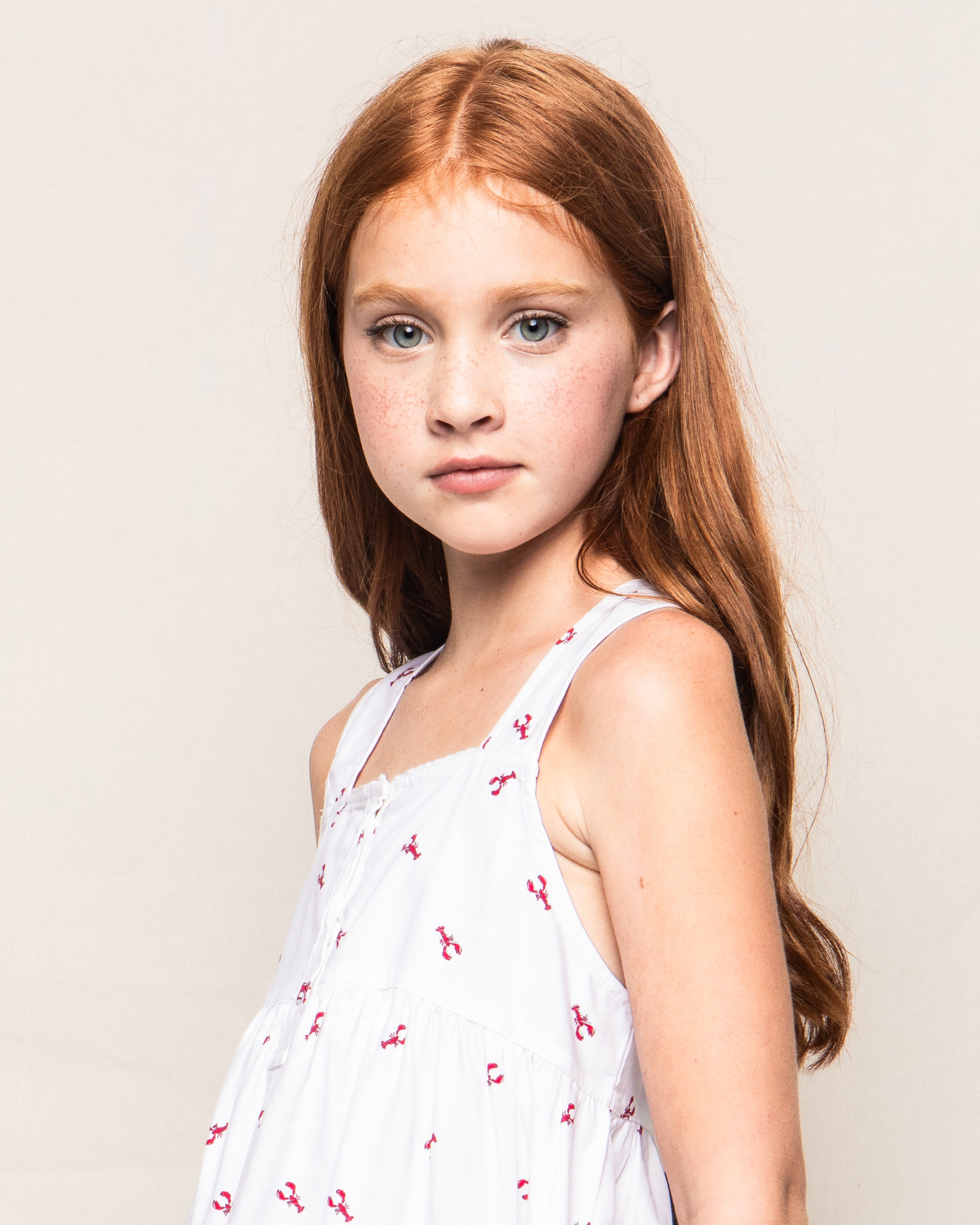 A young girl with long red hair stands calmly against a plain background, wearing Petite Plumes sleeveless Girls Twill Charlotte Nightgown in Brixham Lobsters, exuding an aura of tranquil charm.