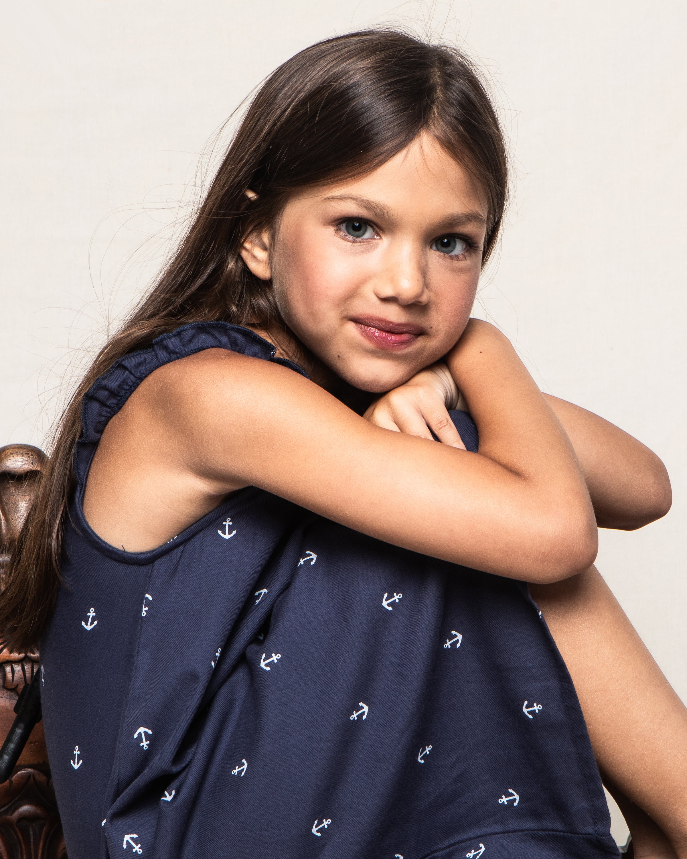 A young girl with long brown hair sits on a chair, smiling at the camera in Petite Plumes Girls Amelie Nightgown in Portsmouth Anchors, perfect for summer days with its lightweight cotton fabric.