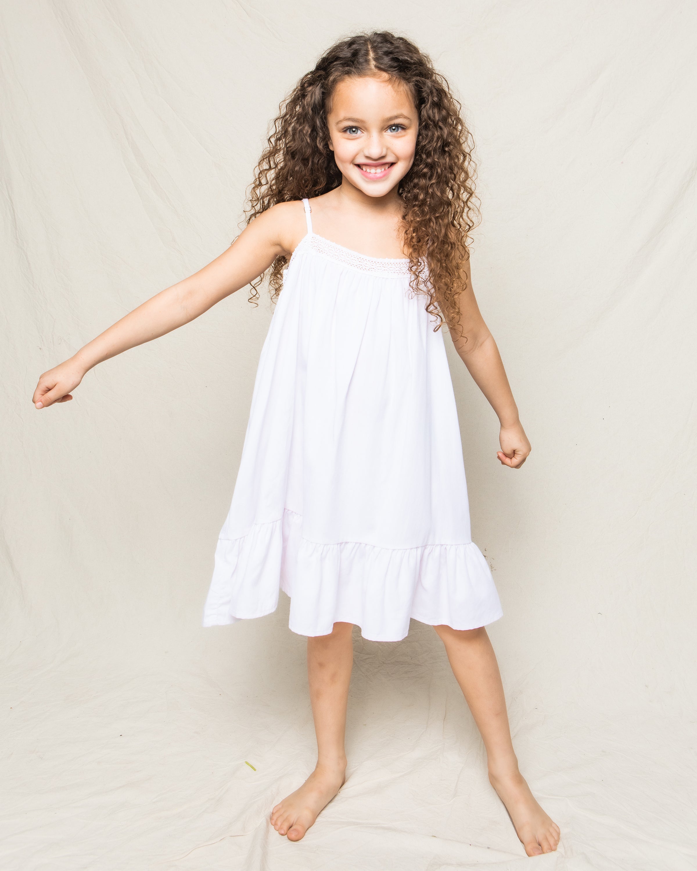 A cheerful young girl with curly hair wears Petite Plumes Girls Twill Lily Nightgown in White, its yarn-dyed cotton adding charm as she smiles barefoot against a light backdrop, her right arm slightly raised.