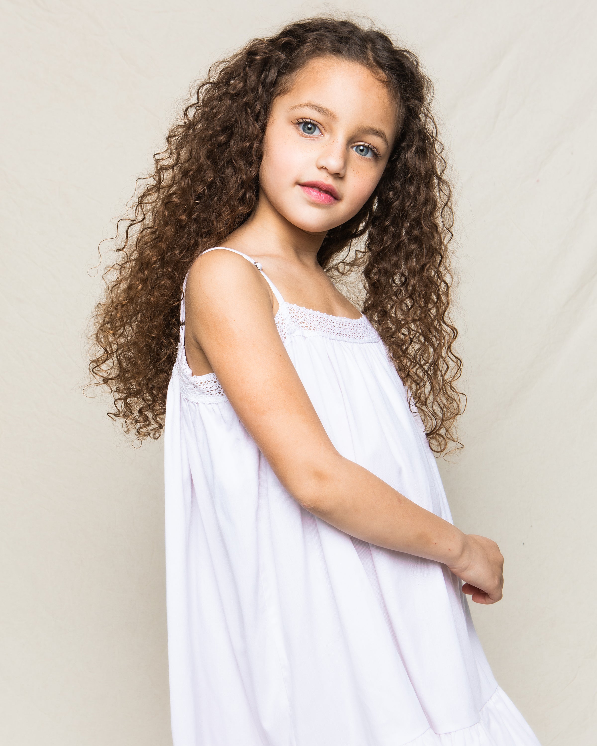 A young girl with long curly hair, wearing a Petite Plume Girls Twill Lily Nightgown in White, stands against a plain light background, slightly smiling at the camera.