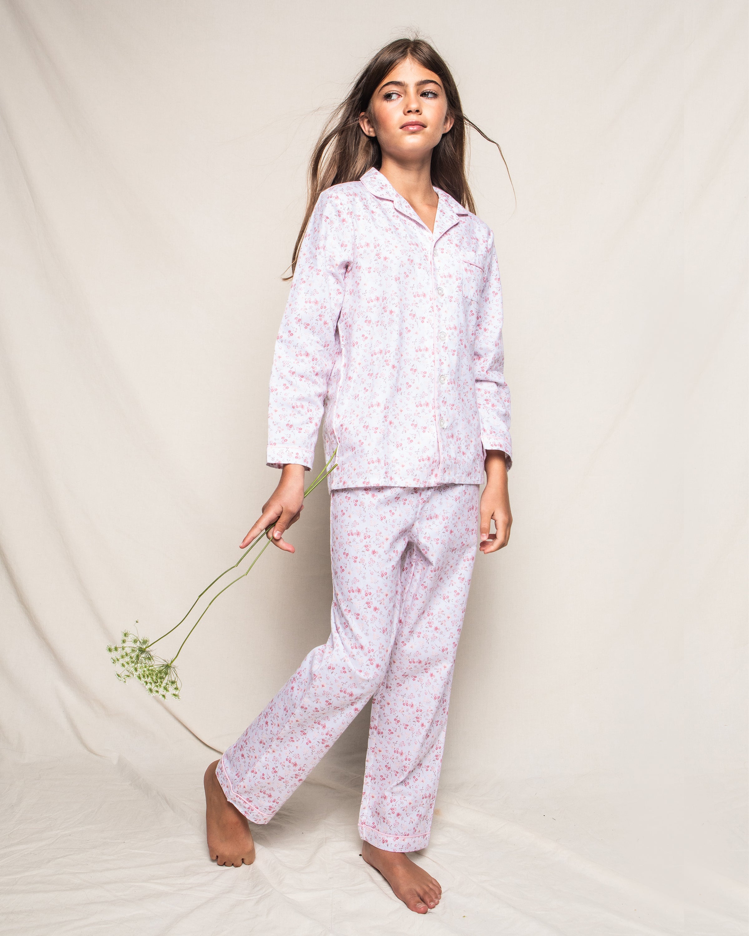 A young girl poses barefoot in the Dorset Floral Kids Twill Pajama Set by Petite Plume, crafted from flame-retardant fibers for safety. She holds delicate white flowers against a neutral backdrop, her hair down loosely.