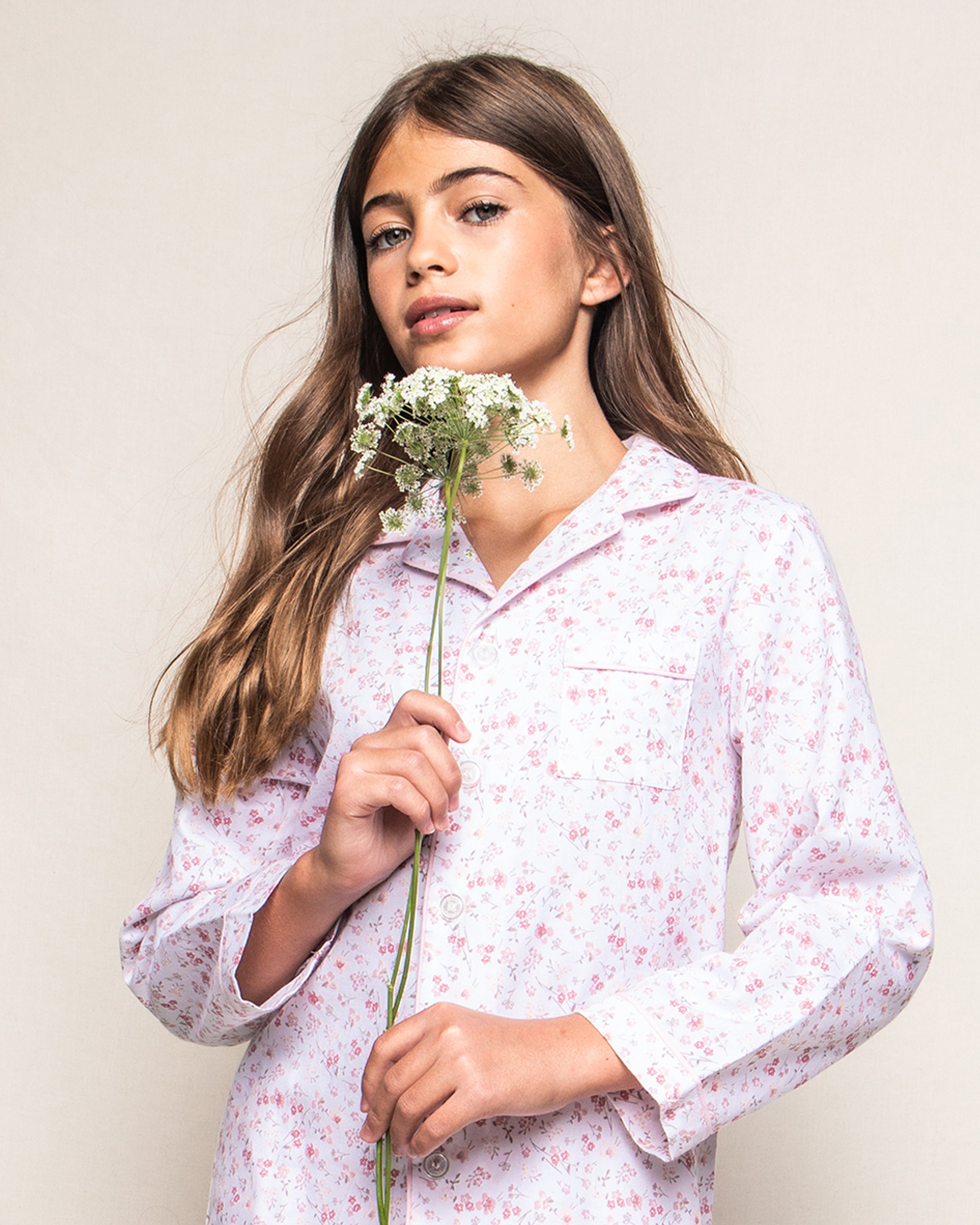 A young girl with long brown hair, holding white flowers, wears the Kids Twill Pajama Set in Dorset Floral by Petite Plume, while standing against a plain background.