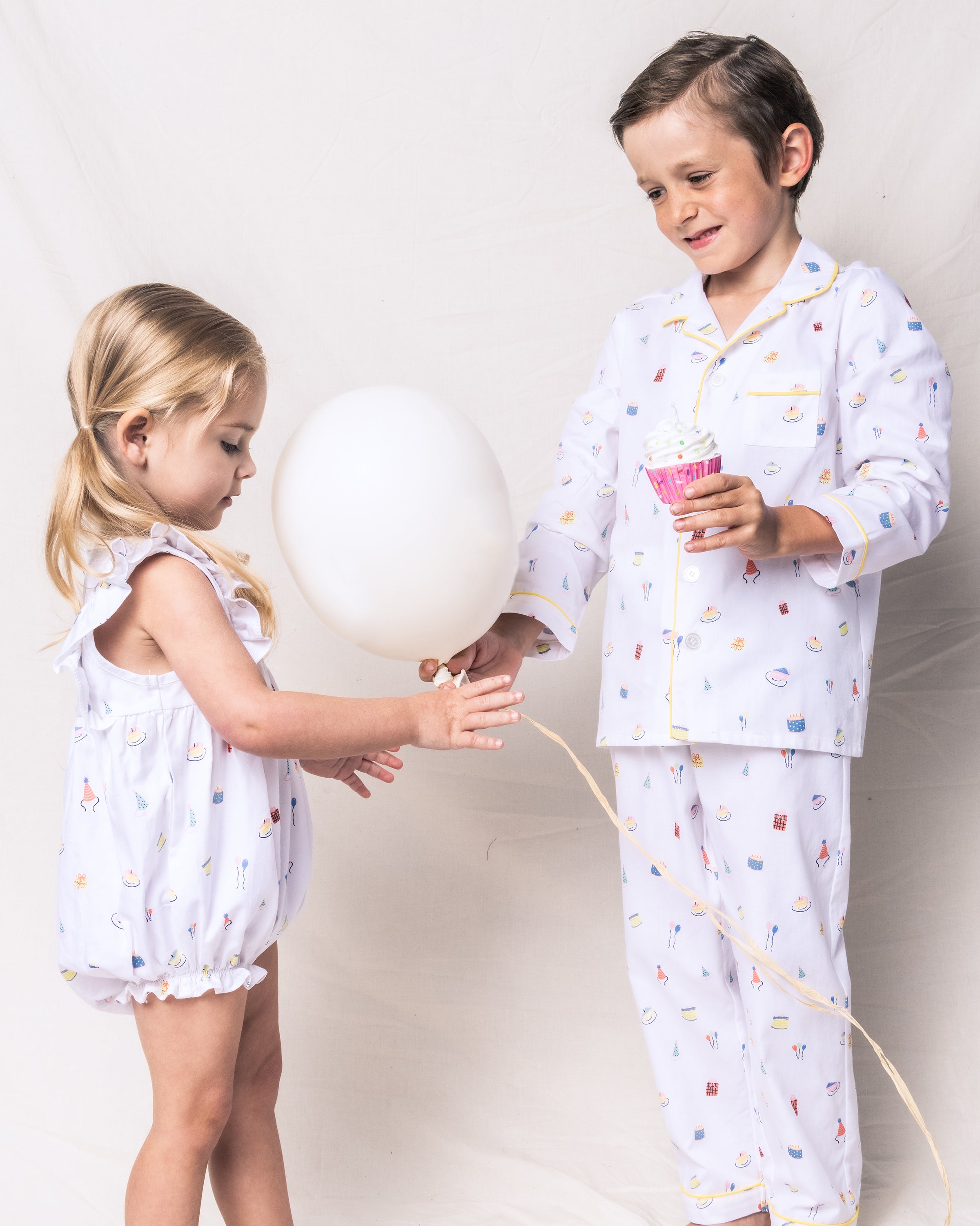 Two kids stand against a light backdrop. The girl in a sleeveless romper with a birthday fantasy print holds a white balloon. The boy, wearing Petite Plumes Kids Twill Pajama Set in Birthday Wishes, holds a pink cupcake liner. Both outfits feature whimsical colorful patterns.