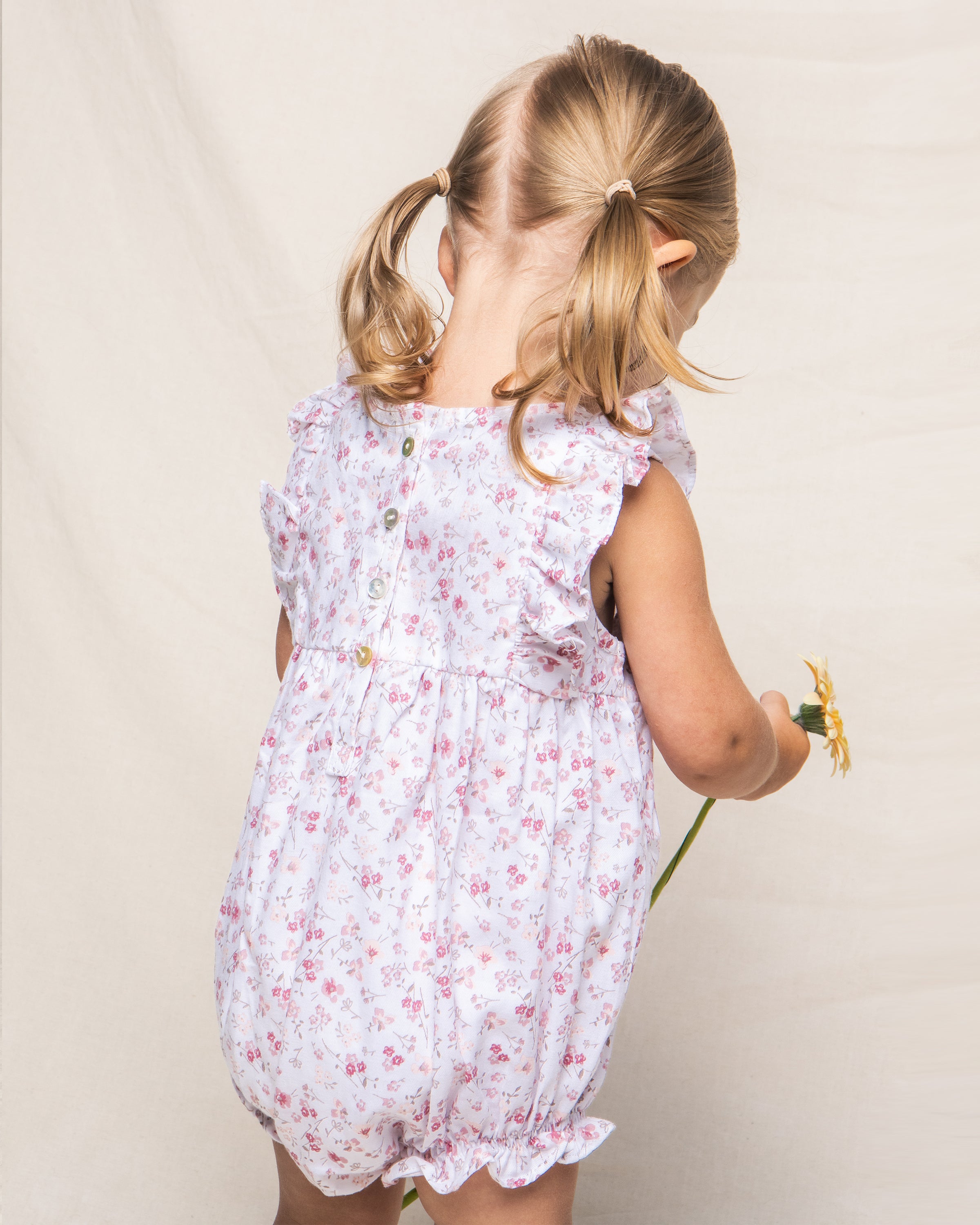 A young girl with pigtails, dressed in Petite Plumes Babys Twill Ruffled Romper in Dorset Floral, is turned away holding a sunflower against a plain light-colored background.