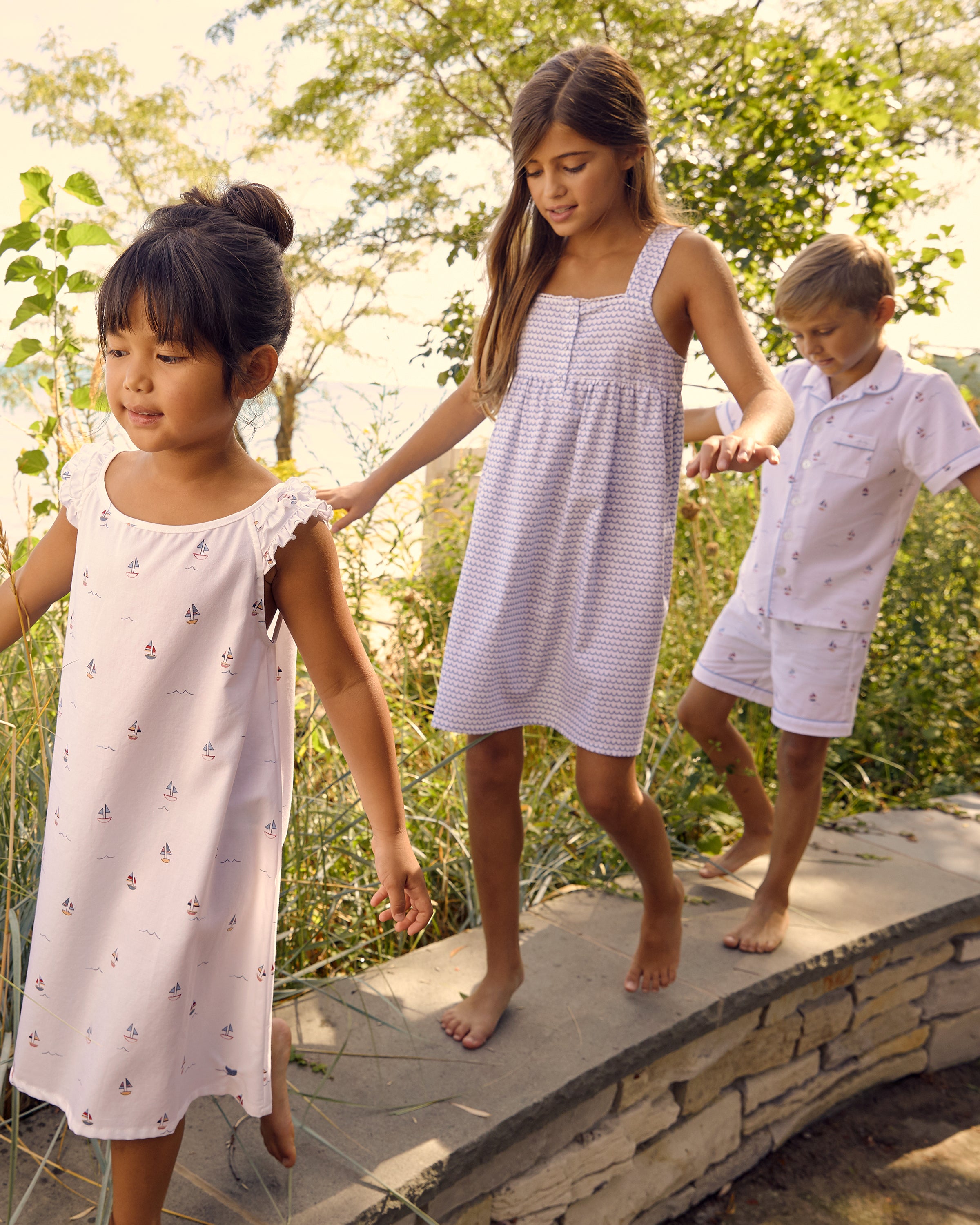 Three children walk barefoot on a stone path by greenery, playfully clutching toy boats. Clad in light summer wear from Petite Plume—two girls in dresses and a boy in the Kids Twill Pajama Short Set in Bateau—the sunny scene is tranquil and idyllic.